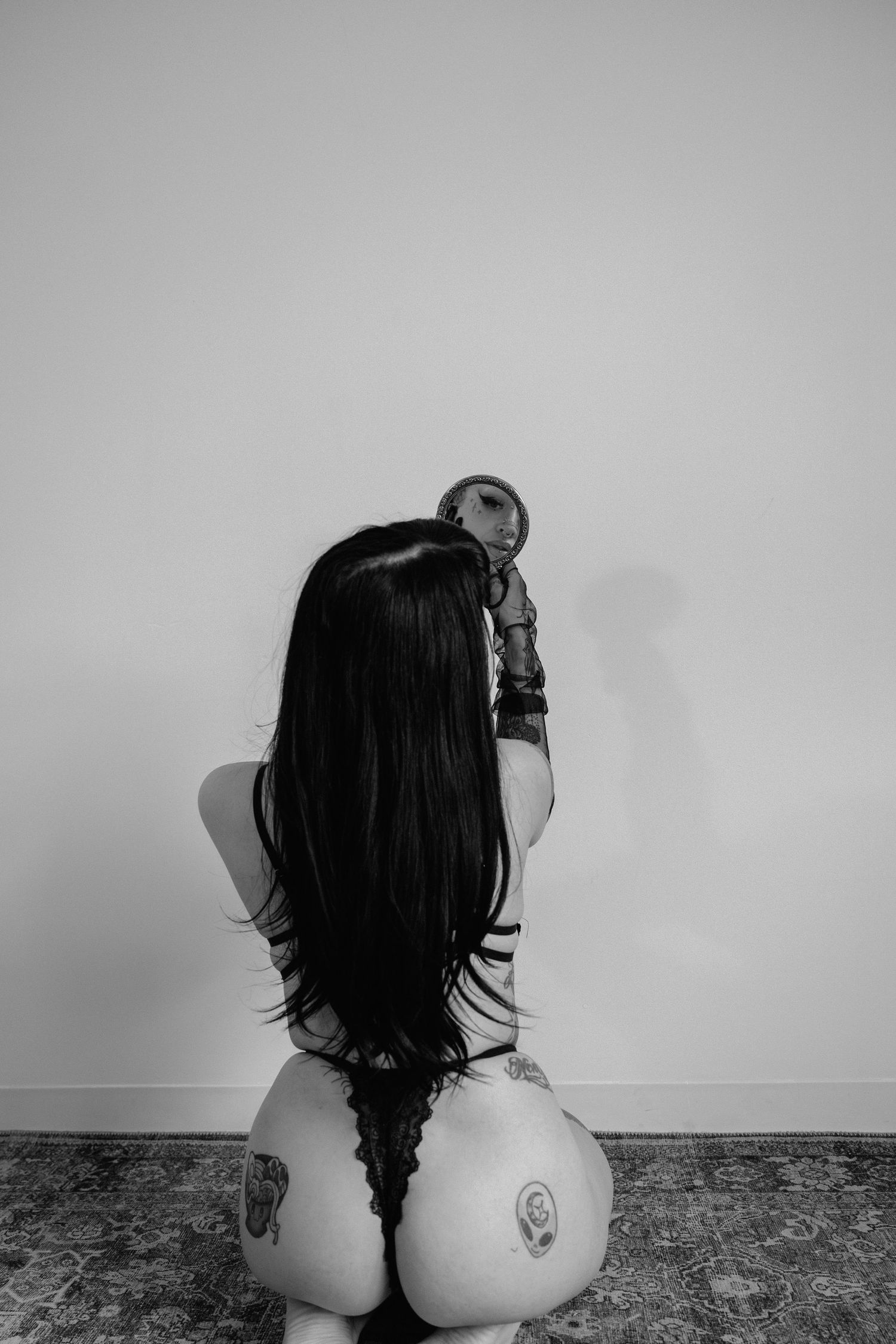 Black and white photo of a tattooed woman in lingerie kneeling, holding a small mirror, viewed from behind.