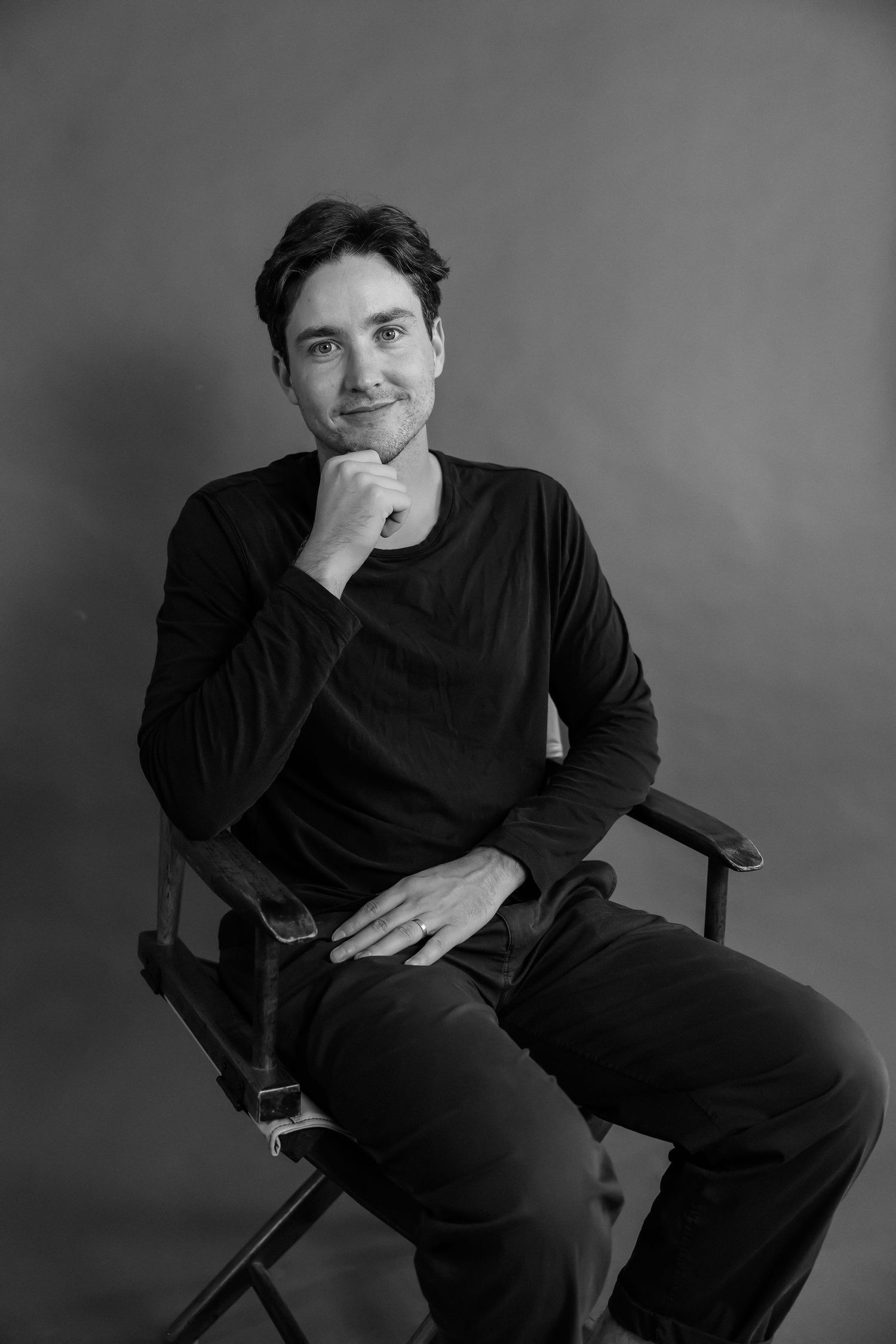 A man in a black sweater sitting in a director's chair, posing thoughtfully in a black and white portrait.