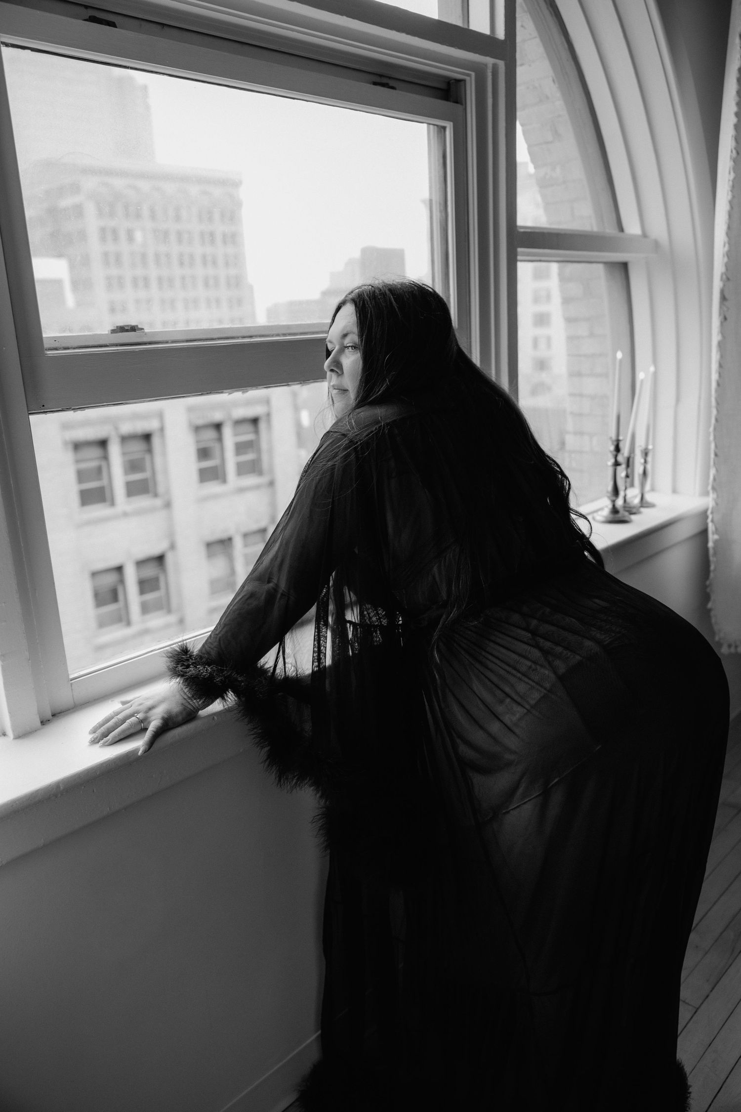 Black and white boudoir photo of a woman in a sheer black robe gazing out a large window overlooking city buildings.