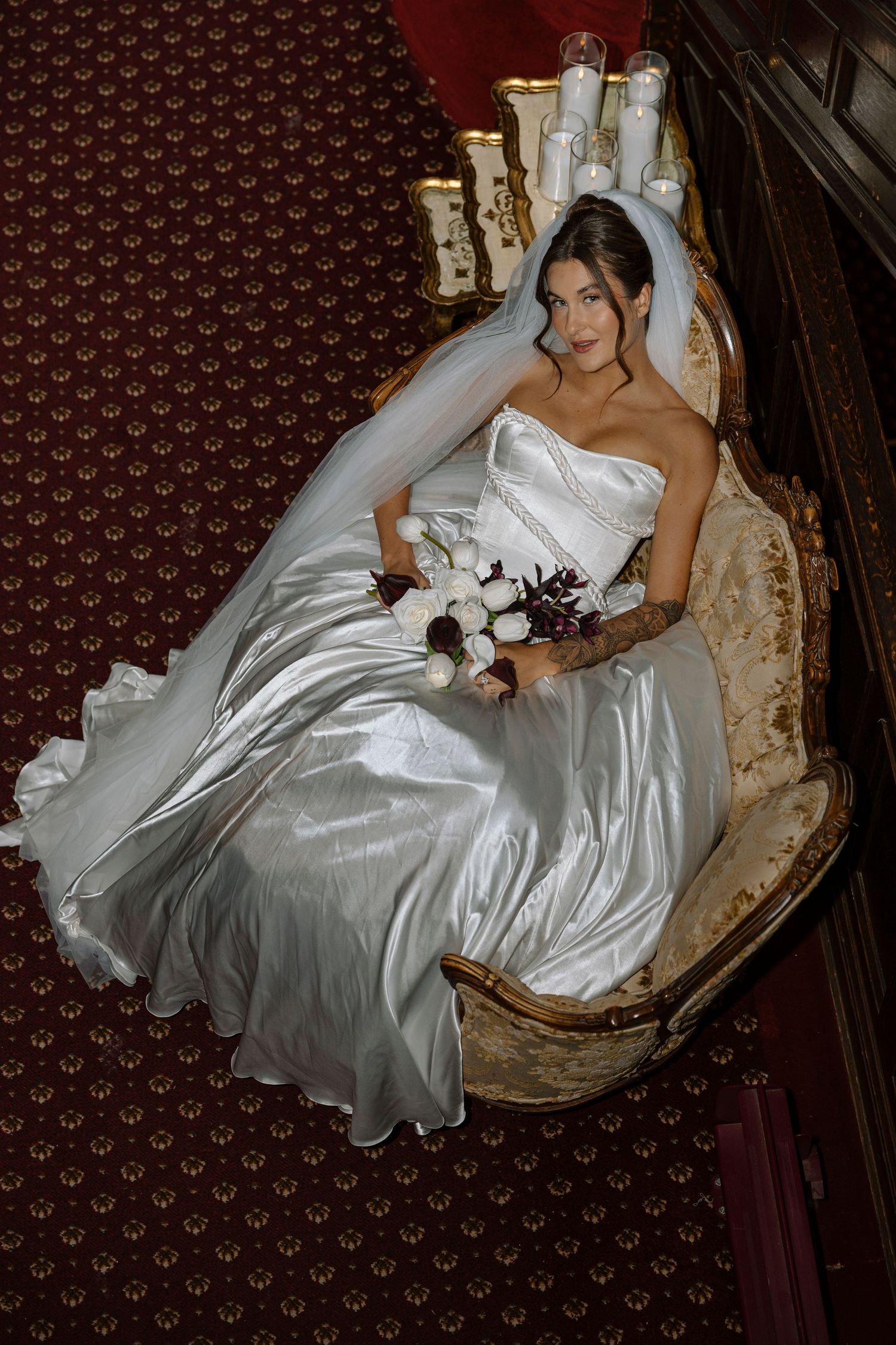 Bride in white satin gown and veil seated on ornate chair, holding bouquet in elegant vintage setting.