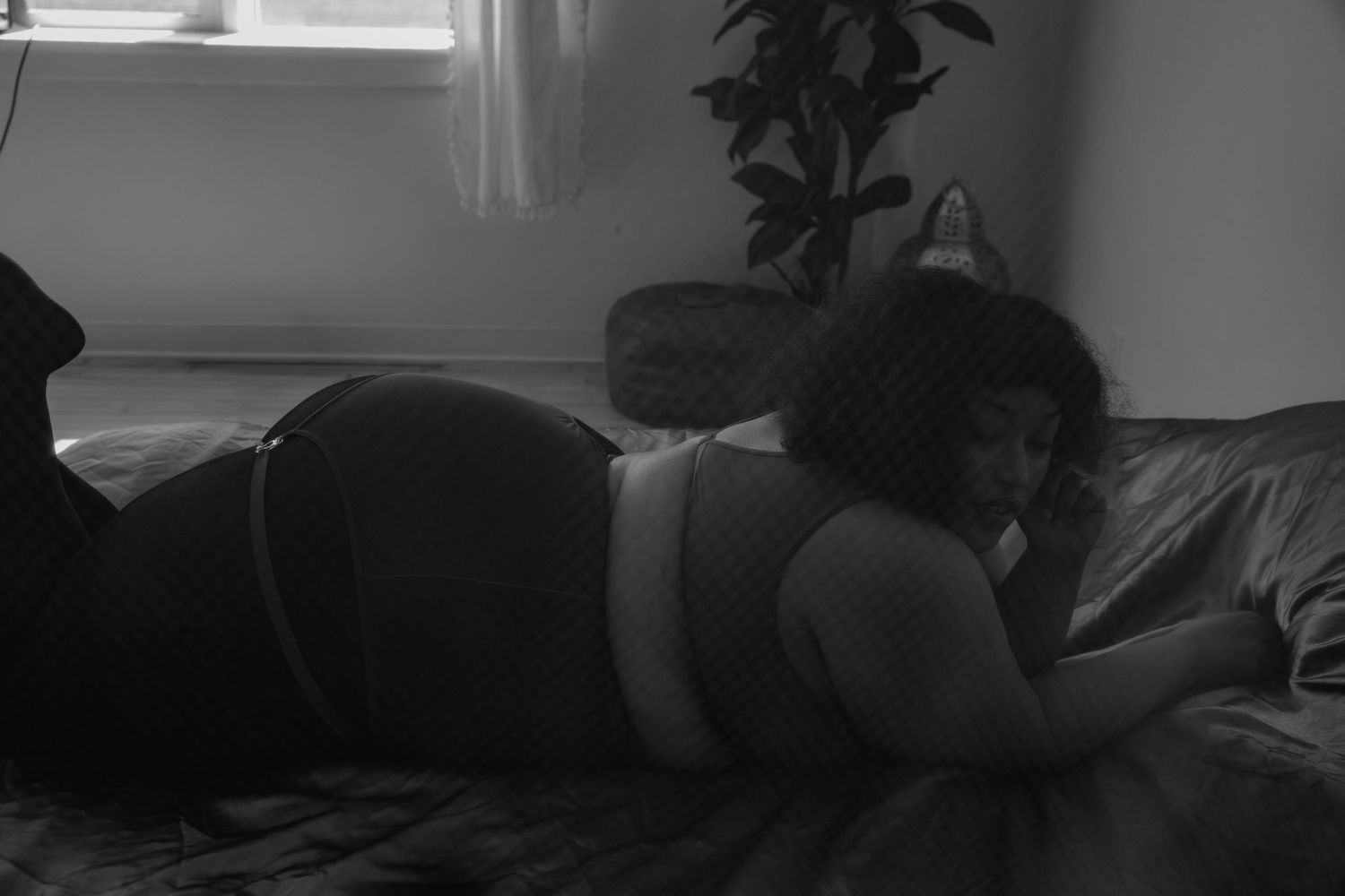 Black and white photo of a woman reclining on a bed in a softly lit bedroom with a plant nearby.