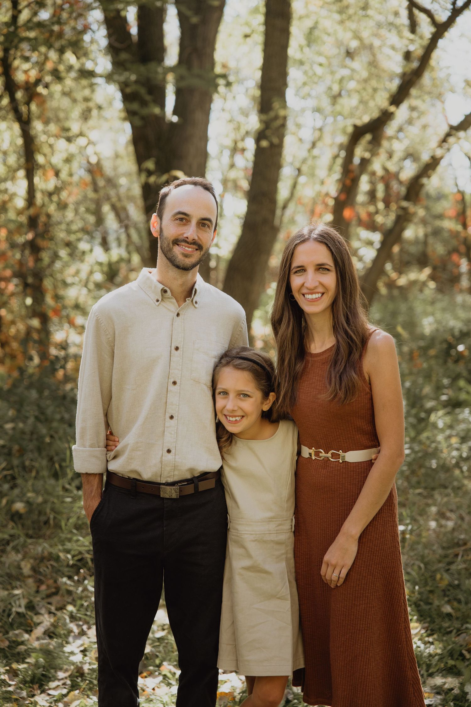 Family of three smiling in autumn forest, man in cream shirt, woman in rust dress, young girl between them.