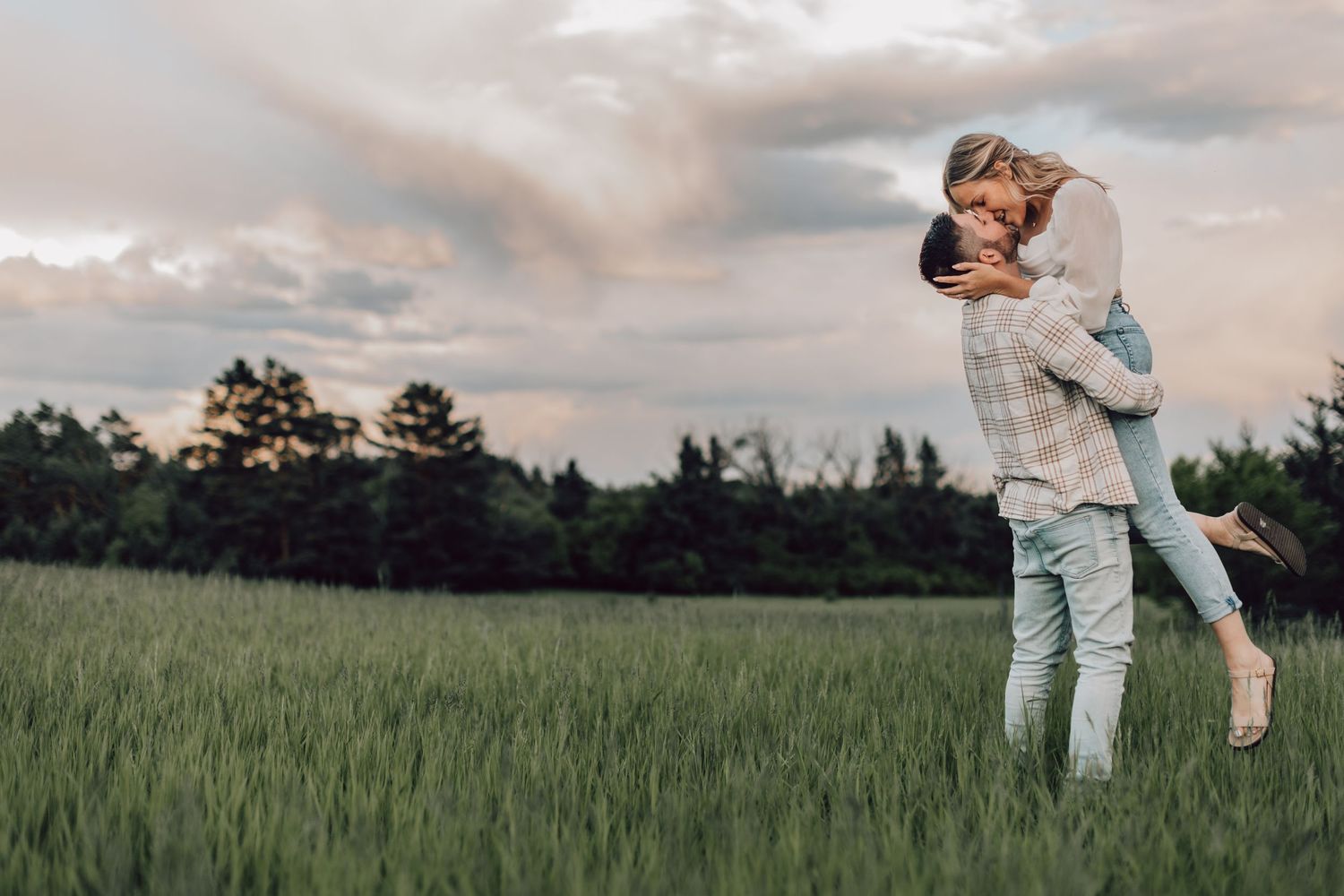 Couple embracing and lifting each other joyfully in a lush green field under a dramatic cloudy sky.
