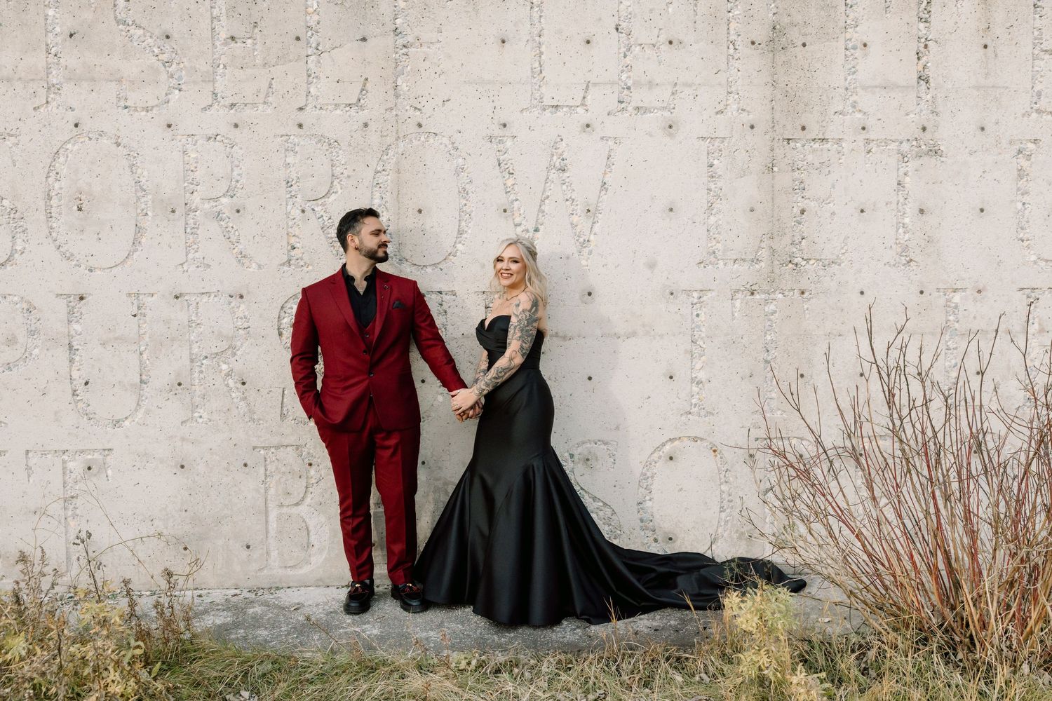 Couple posing by concrete wall, man in red suit, woman in black mermaid gown, holding hands outdoors.