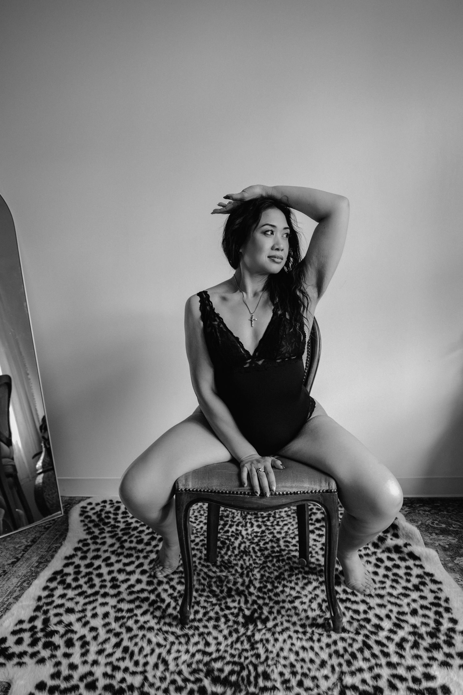 Black and white boudoir photo of a woman in a bodysuit sitting on a stool on a leopard print rug.
