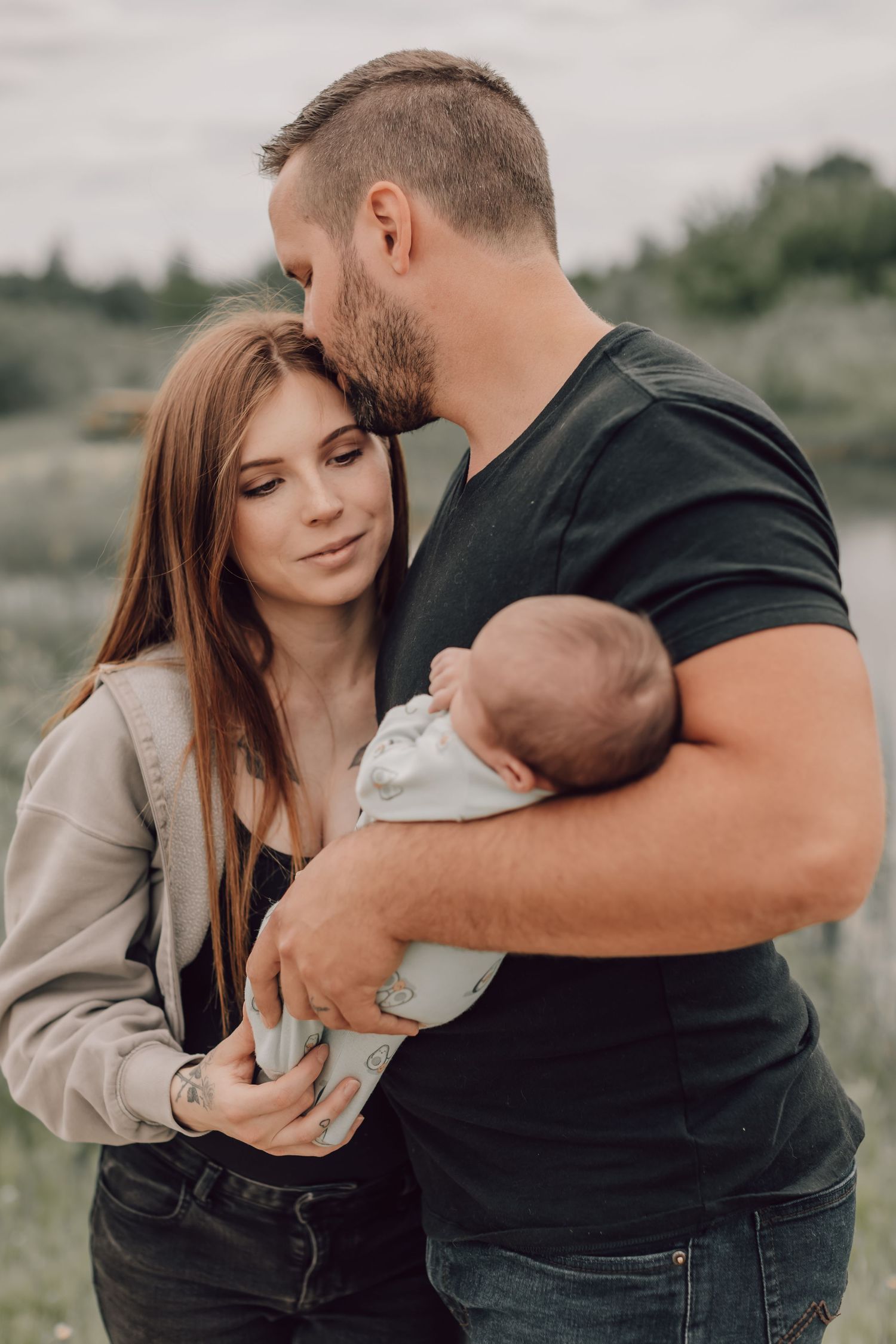 Young couple tenderly embracing outdoors while father gently holds their newborn baby.