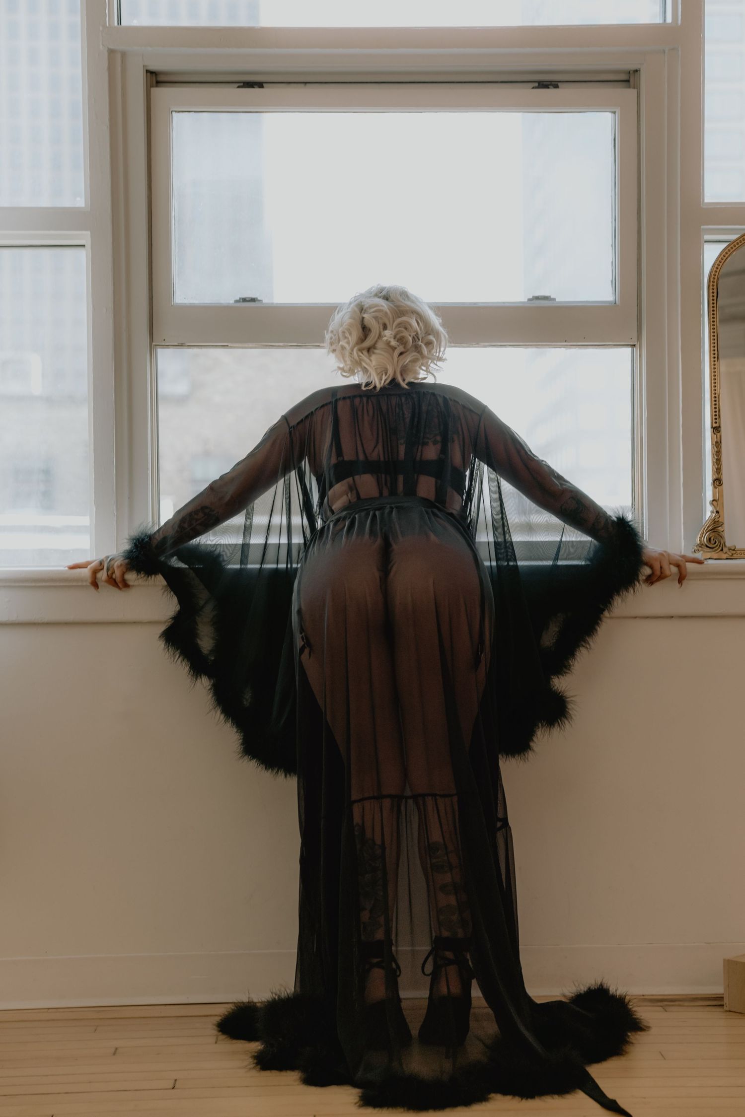 Woman in sheer black maxi robe with feather trim stands facing window in elegant boudoir photography.