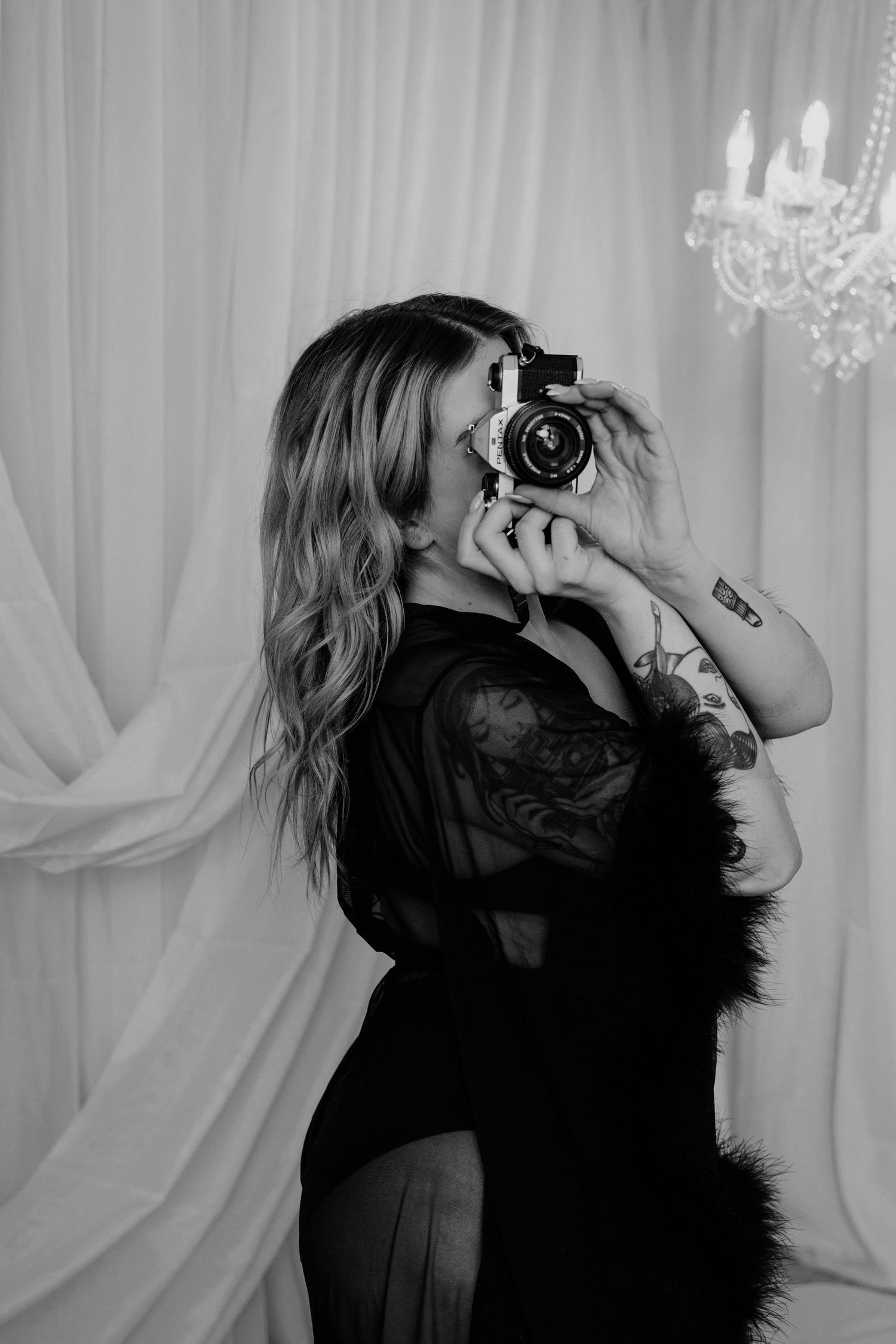 Black and white photo of tattooed woman in black robe holding vintage camera, chandelier backdrop.