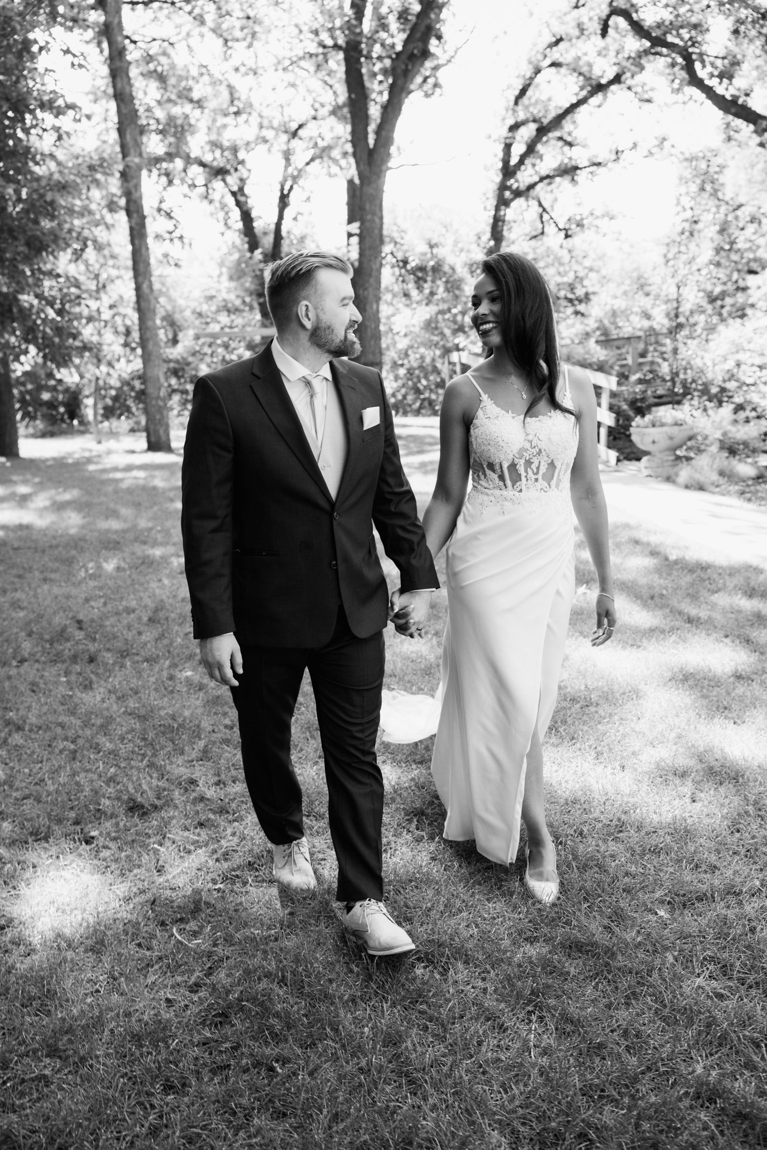 Black and white photo of a smiling couple in wedding attire holding hands and walking through a park.