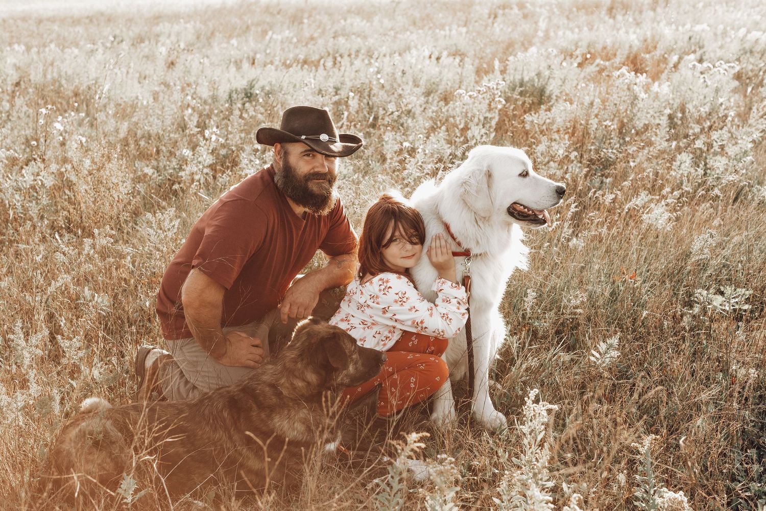 Bearded man in cowboy hat sitting in a wildflower field with a child and two dogs.