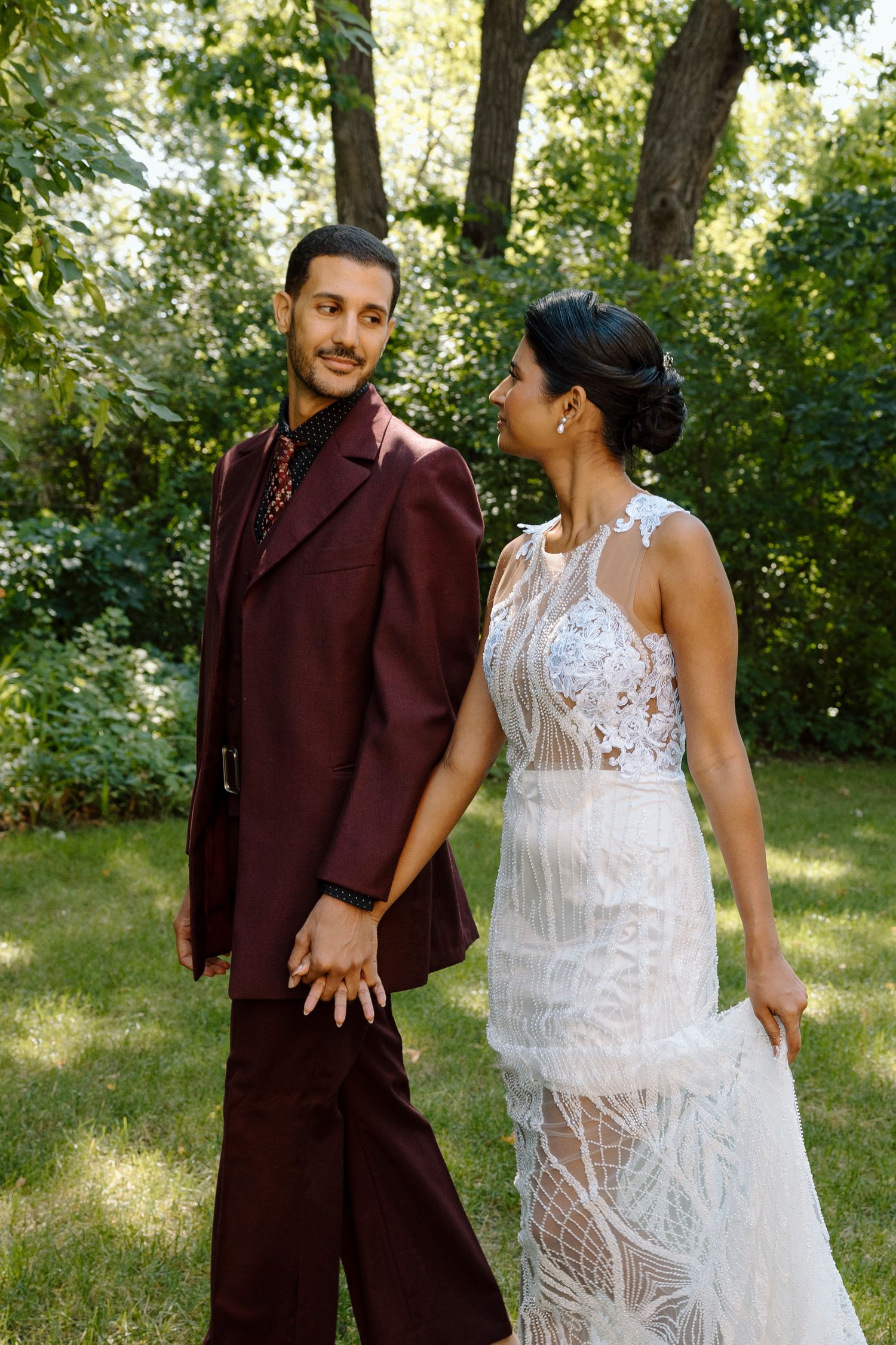 Couple in wedding attire holding hands outdoors, groom in burgundy suit, bride in white lace gown, green trees background.