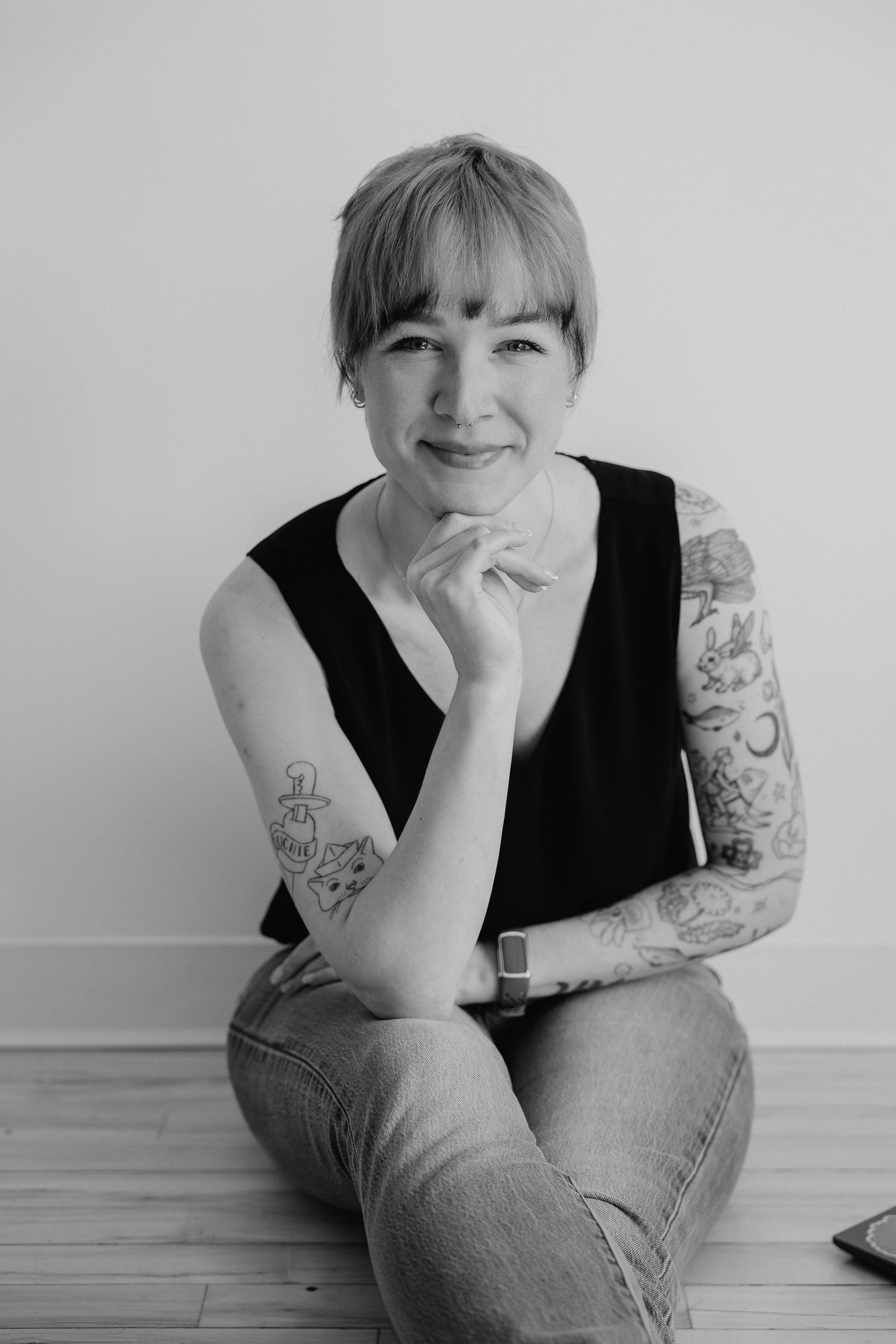 Black and white portrait of a tattooed woman sitting on a floor, smiling warmly.