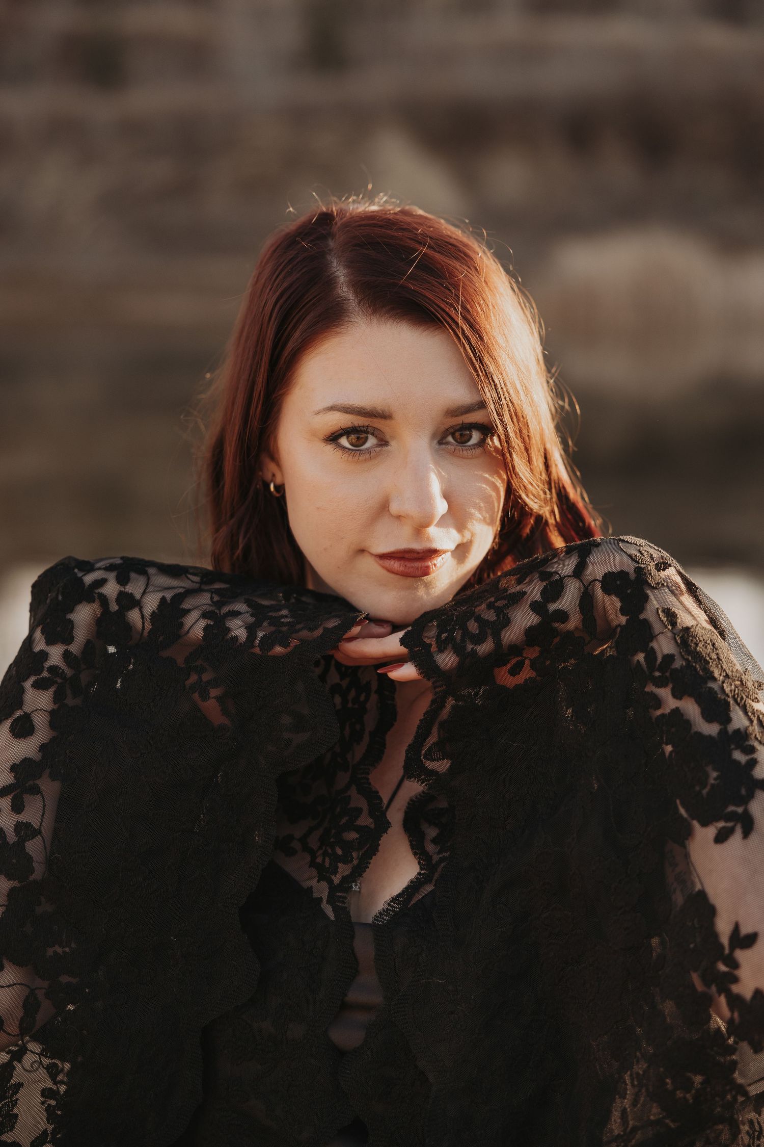 Portrait of a red-haired woman in black lace wrap gazing at camera in warm golden light.
