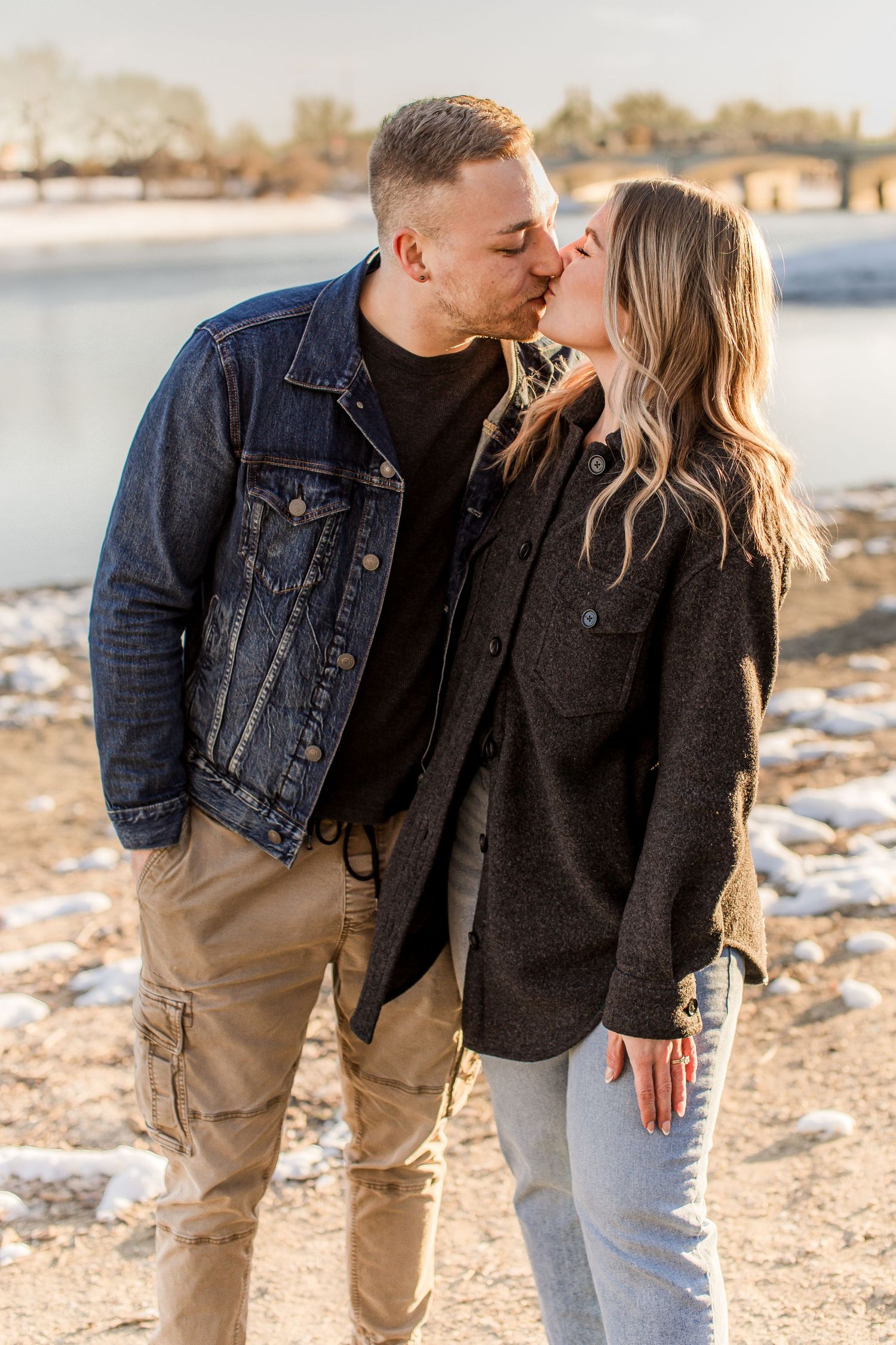 Couple sharing a tender kiss outdoors near a frozen lakeside on a sunny winter day.