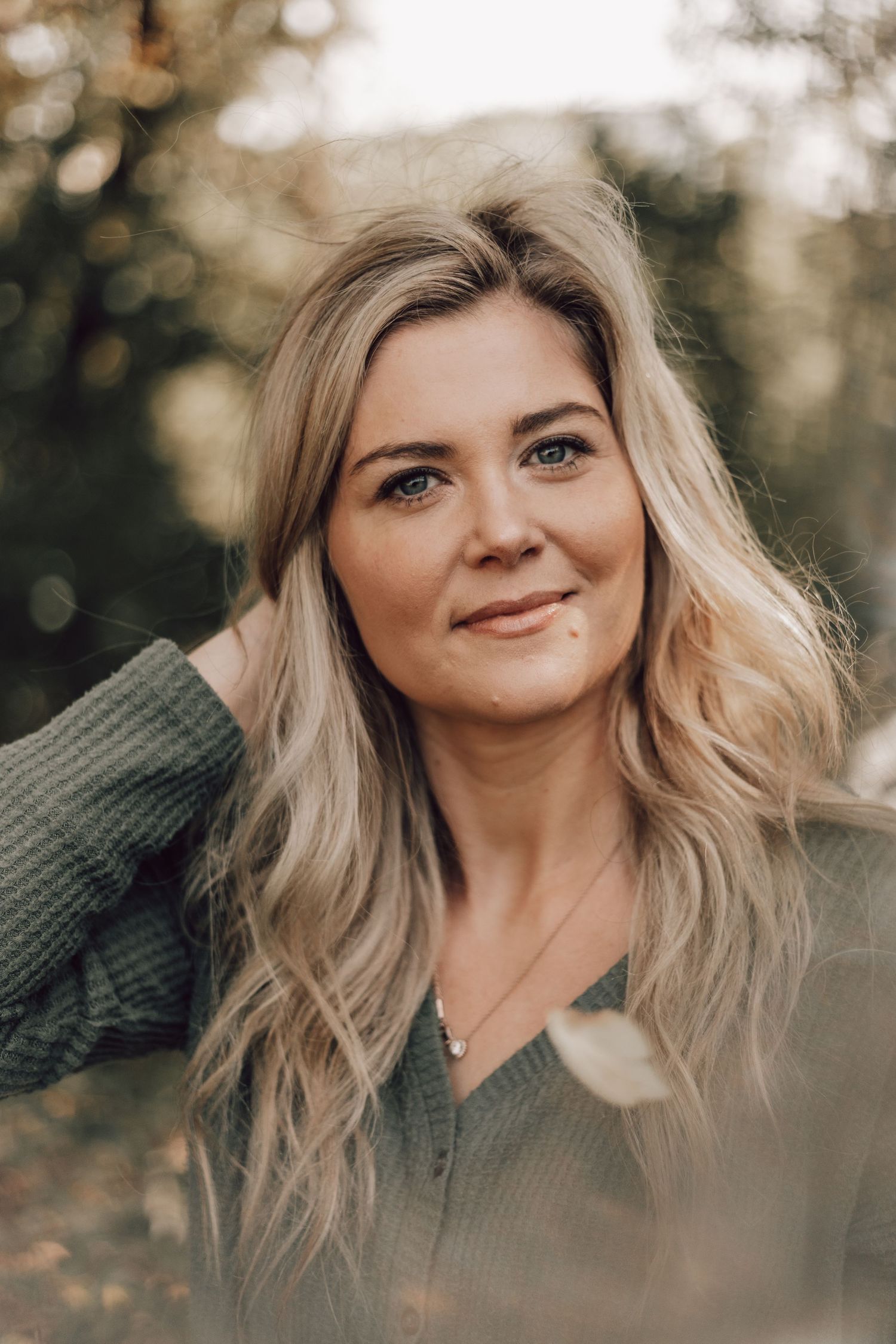 Blonde woman in green sweater smiling softly while touching her hair in an outdoor setting.