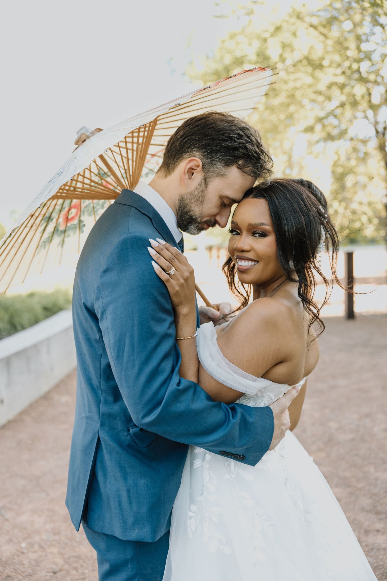 Interracial couple embraces under a decorative parasol, bride in off-shoulder gown smiling joyfully.