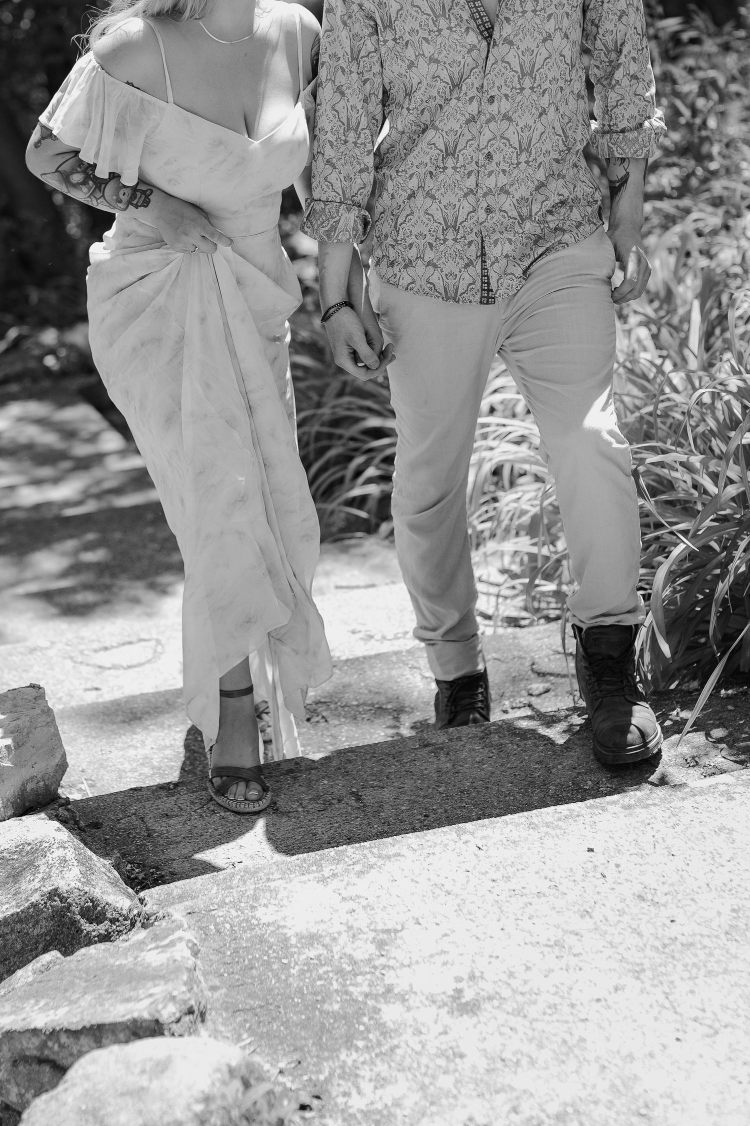 Black and white photo of a couple holding hands while walking on rocky path in a lush garden.