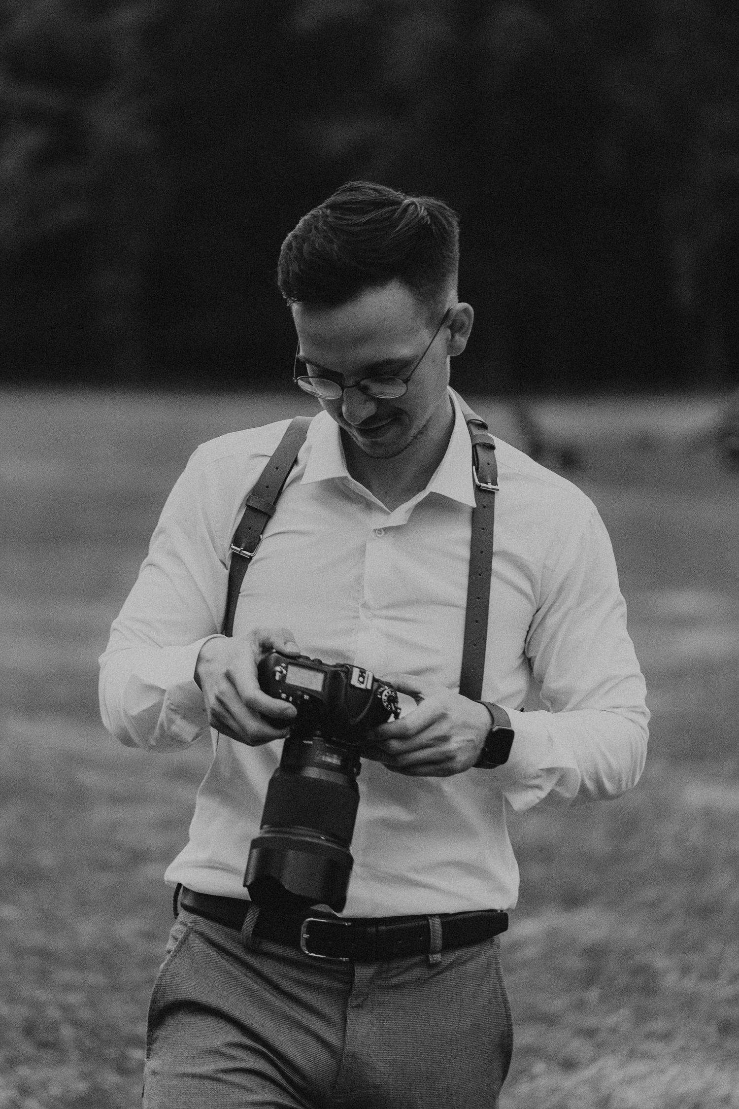Black and white portrait of photographer in suspenders examining camera outdoors.