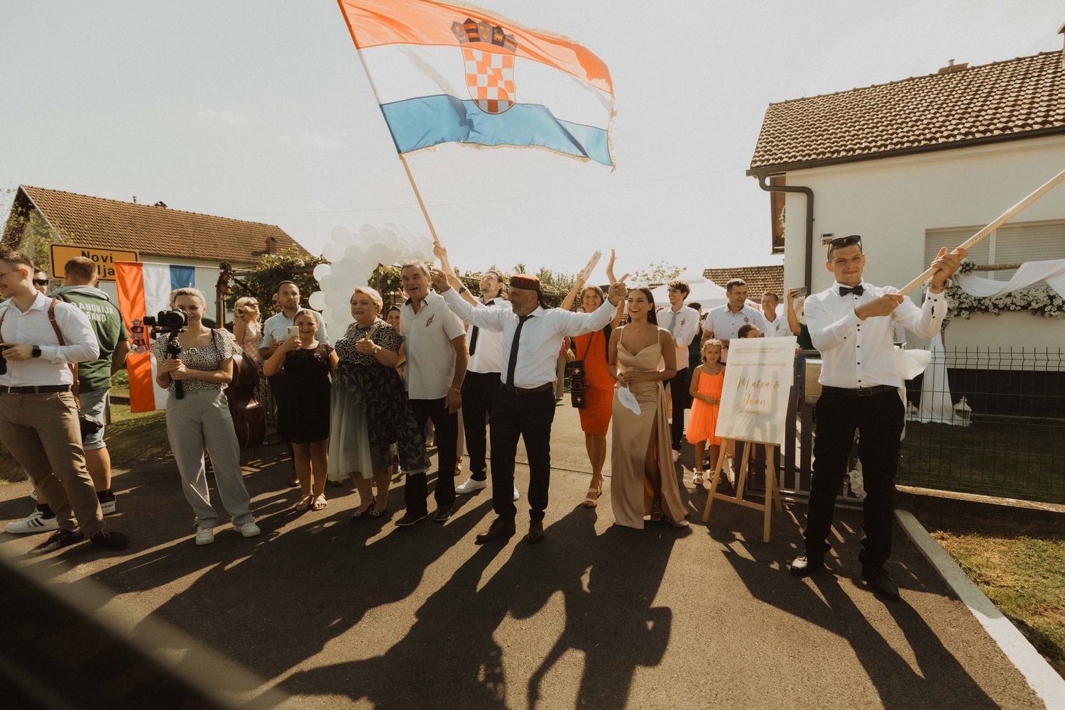 Joyful crowd celebrates outdoors with an orange and blue flag against houses in golden sunlight.