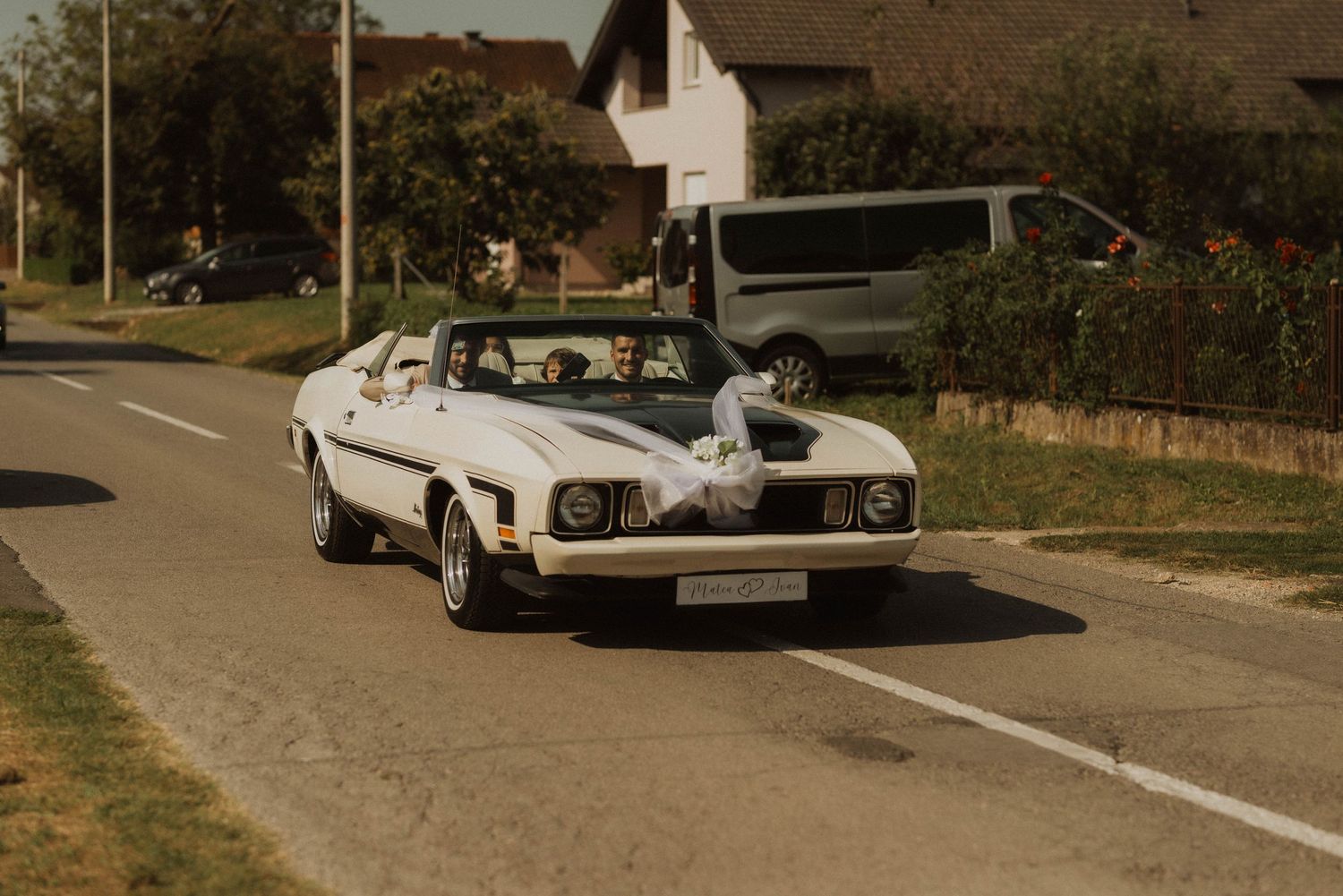 A classic white vintage car decorated with wedding ribbons driving down a suburban street.