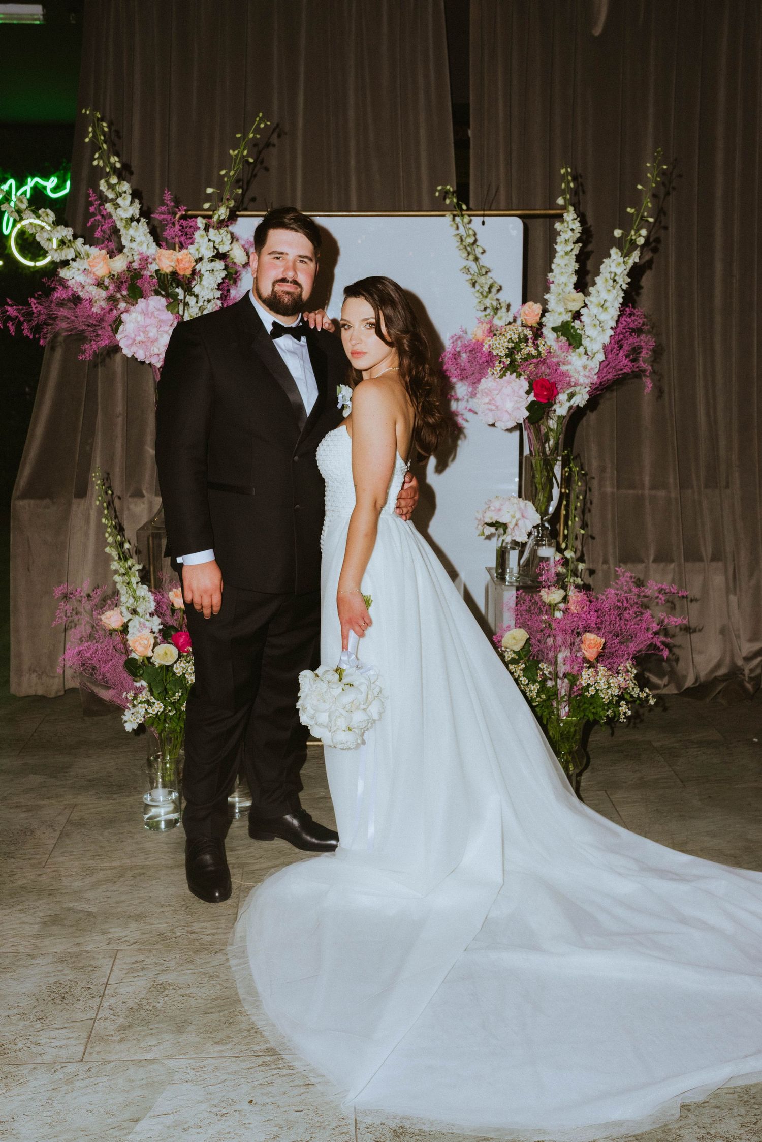 Wedding couple poses before floral arch with pink and purple blooms in elegant venue.