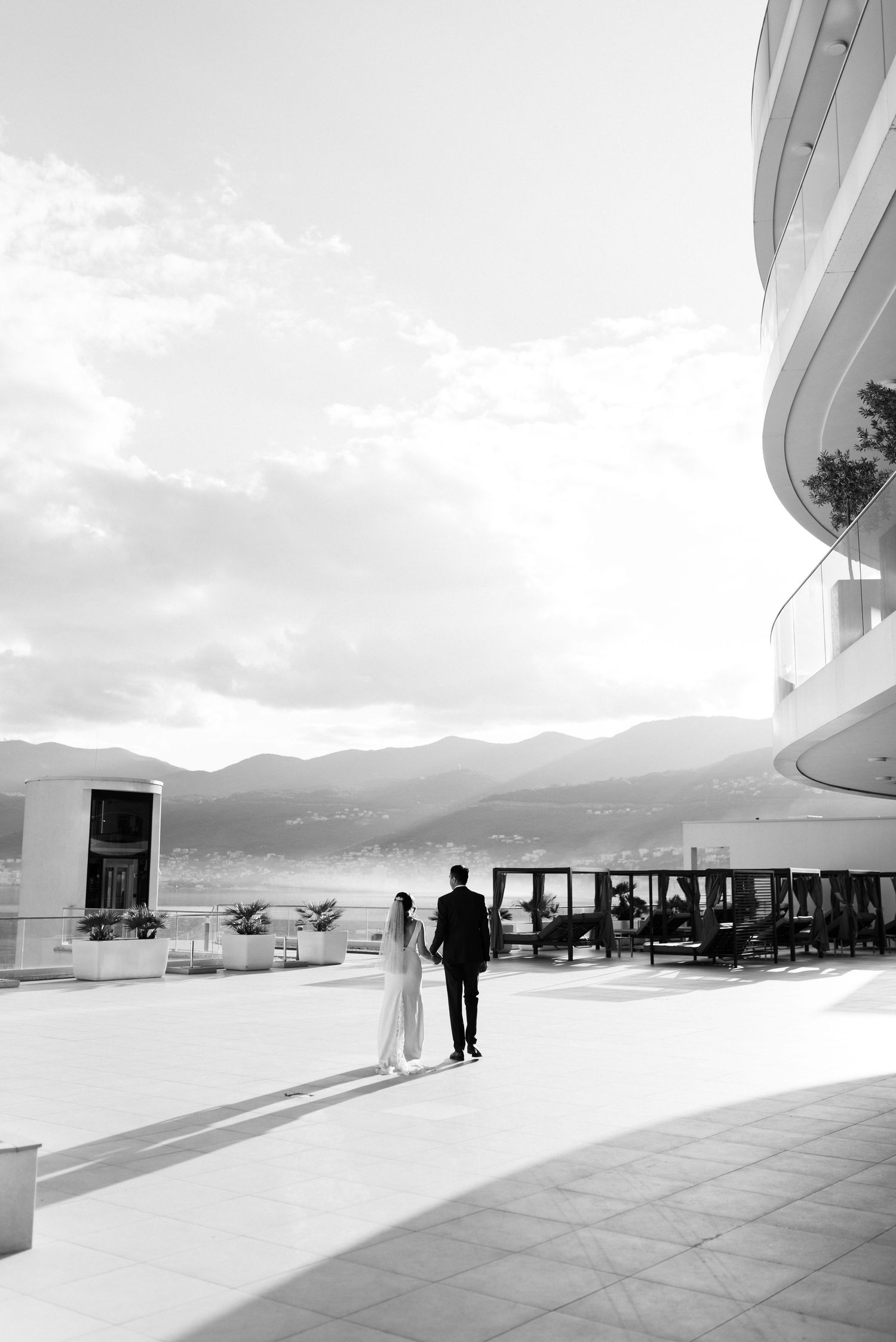 Black and white wedding photo of couple walking on modern terrace overlooking mountain vista.