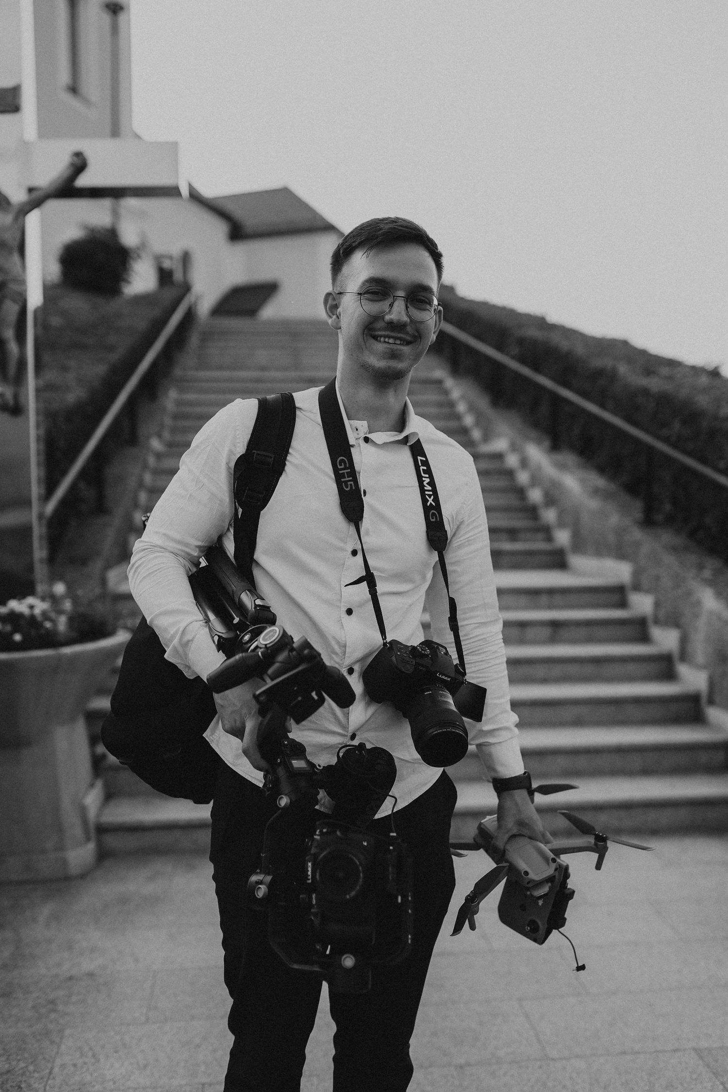 Professional photographer carrying multiple cameras standing on concrete steps in black and white.