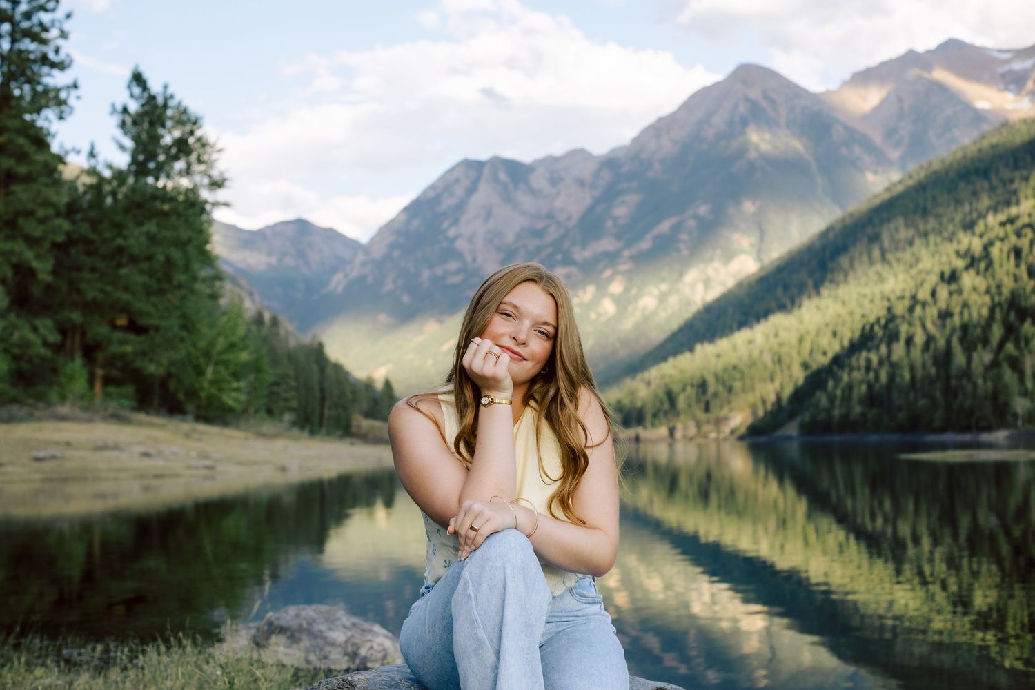 Young woman sitting by a reflective mountain lake surrounded by pine trees and peaks, smiling warmly in golden hour light.