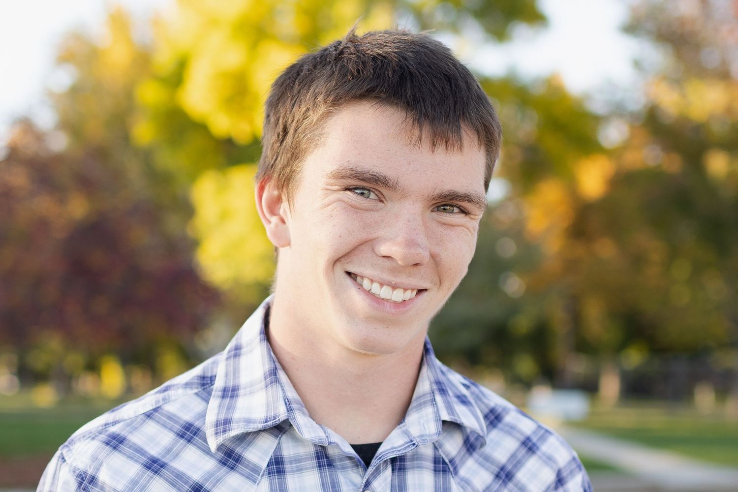 Smiling young man in plaid shirt outdoors with colorful autumn trees in background.
