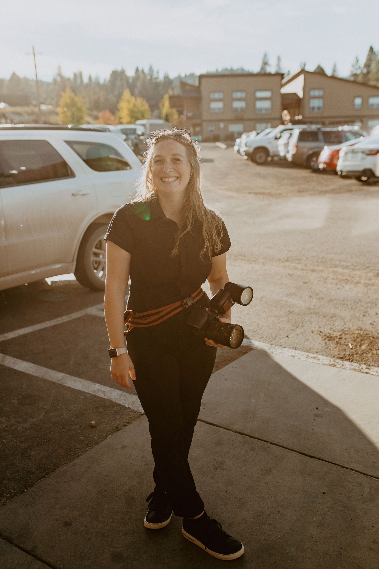 Smiling female photographer holding a camera in a sunlit parking lot, wearing all black with an orange camera strap.