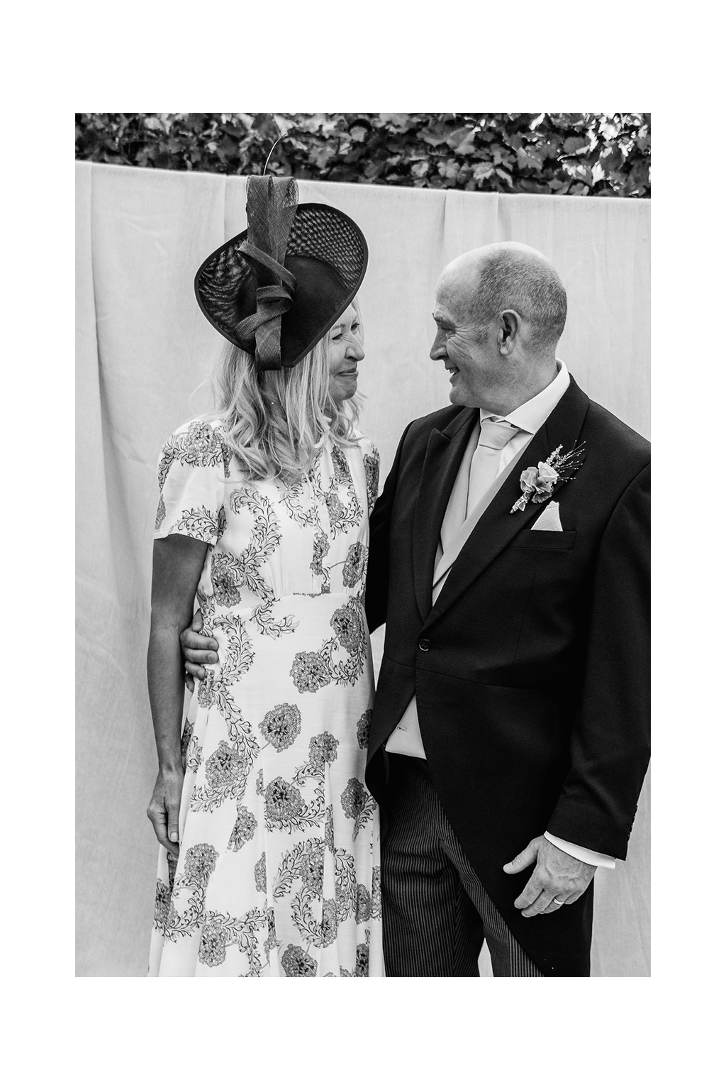 Black and white photo of a smiling woman in a floral dress and fascinator embracing a man in a morning suit with a boutonniere at a wedding.