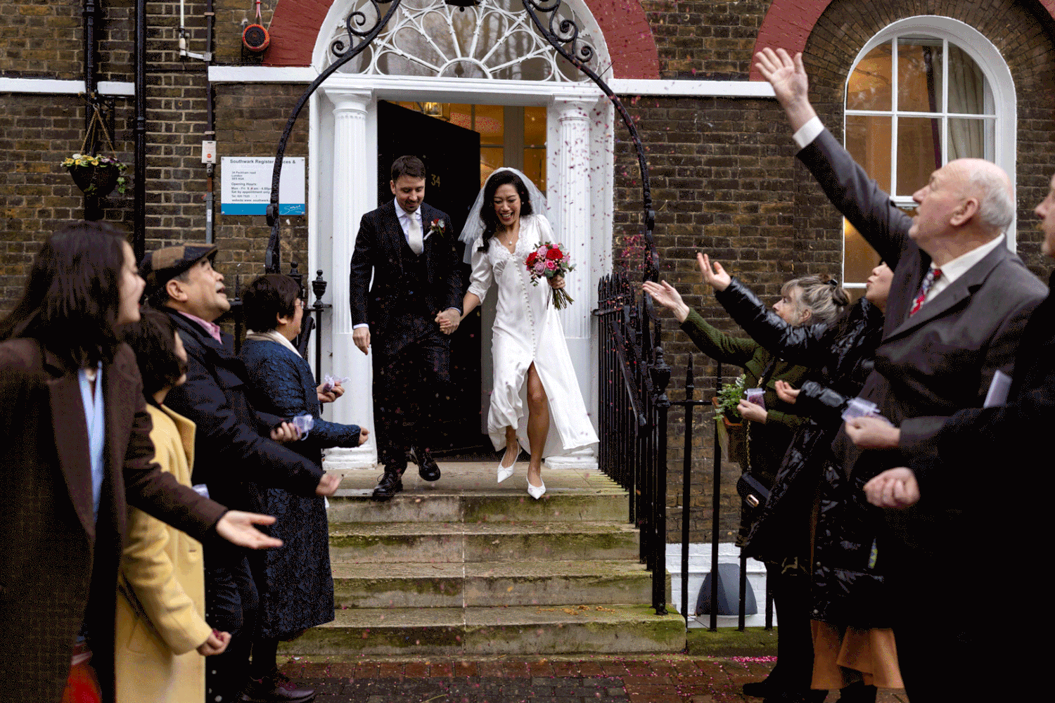 Wedding party members pose on brick steps during outdoor celebration with guests taking photos.