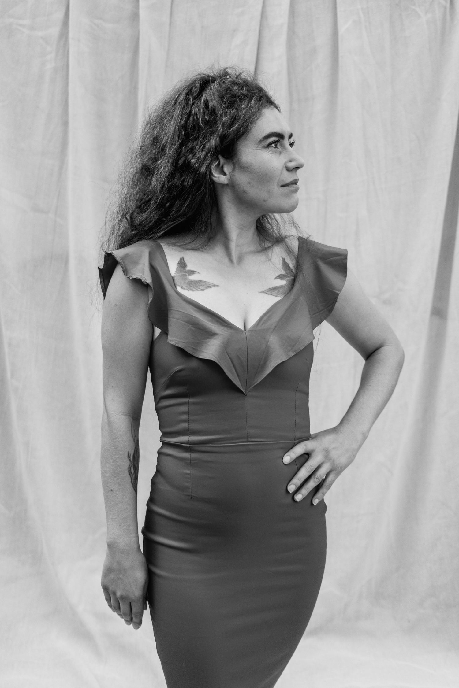 Black and white portrait of a woman with long curly hair in a fitted dress with a ruffled neckline, looking aside against a draped backdrop.