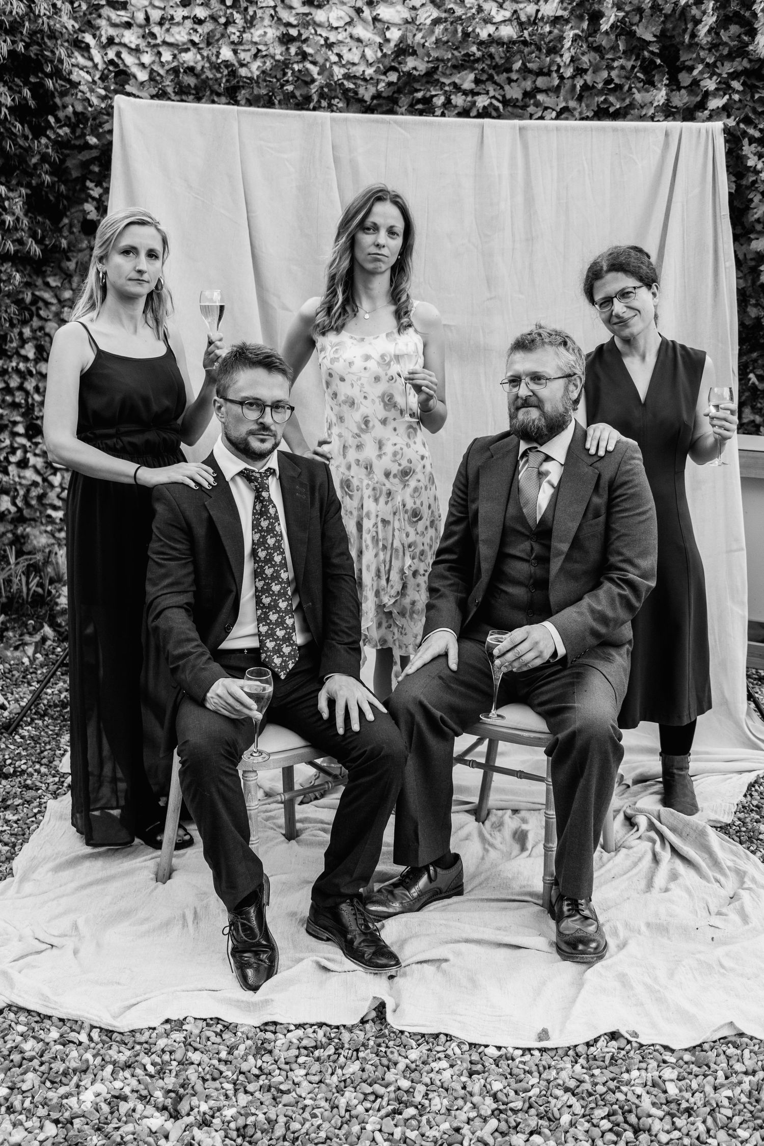 Black and white portrait of two seated men in suits and three standing women holding wine glasses before a draped backdrop outdoors.