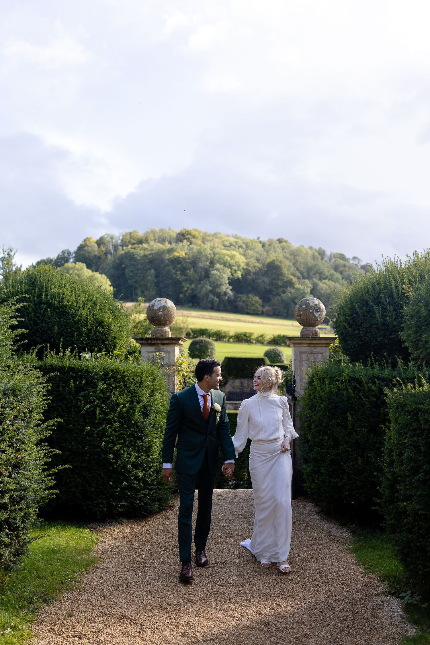 A couple walks down a garden path surrounded by manicured hedges and scenic countryside views on a sunny day.