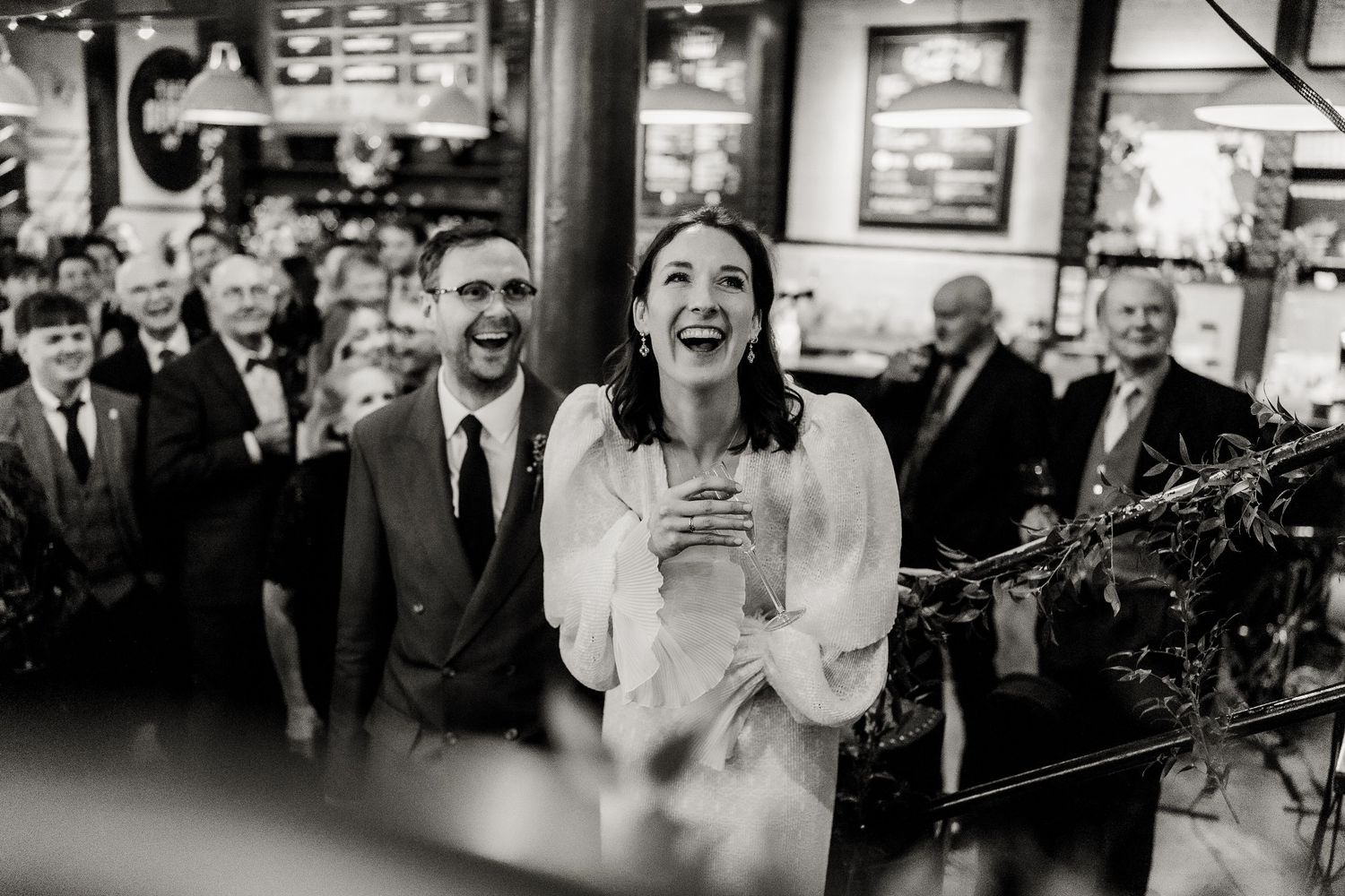 Black and white photo of a laughing bride holding a drink surrounded by wedding guests in a venue.
