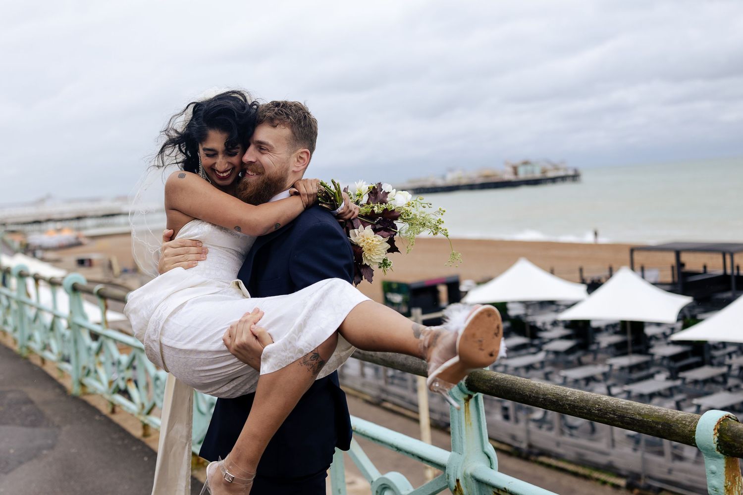 A couple shares an intimate embrace against a scenic waterfront backdrop with pier and boats in the distance.