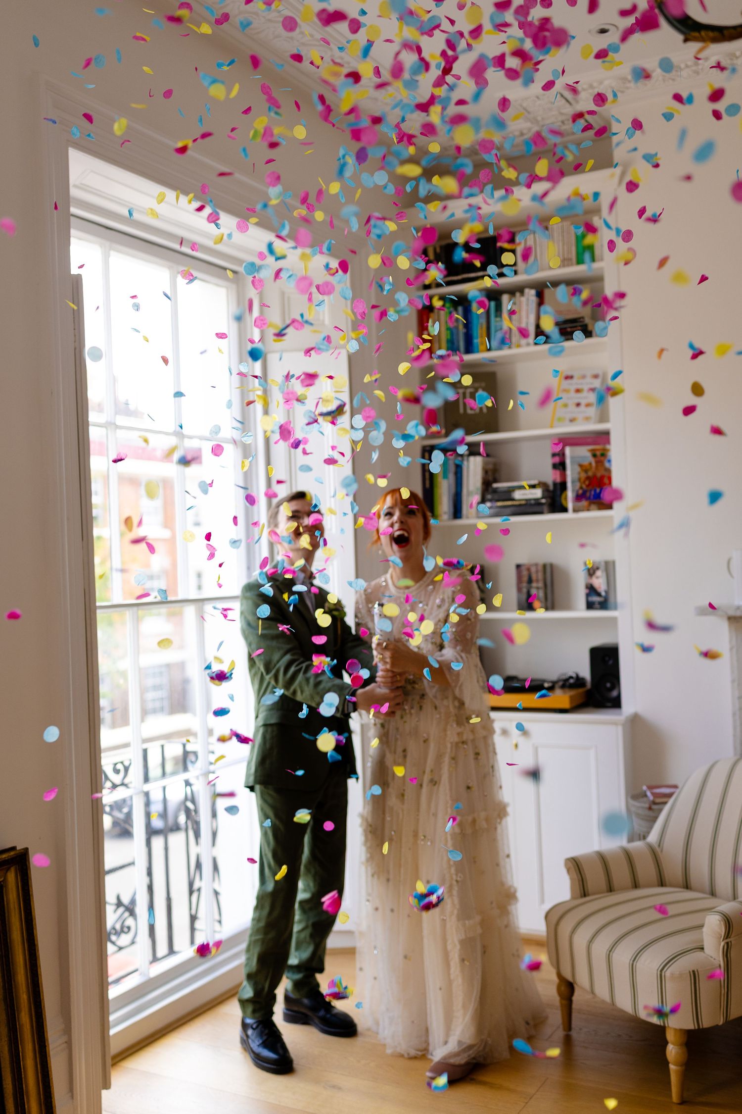 Colorful confetti falls around a couple in formal attire standing by a bright window and built-in shelving.