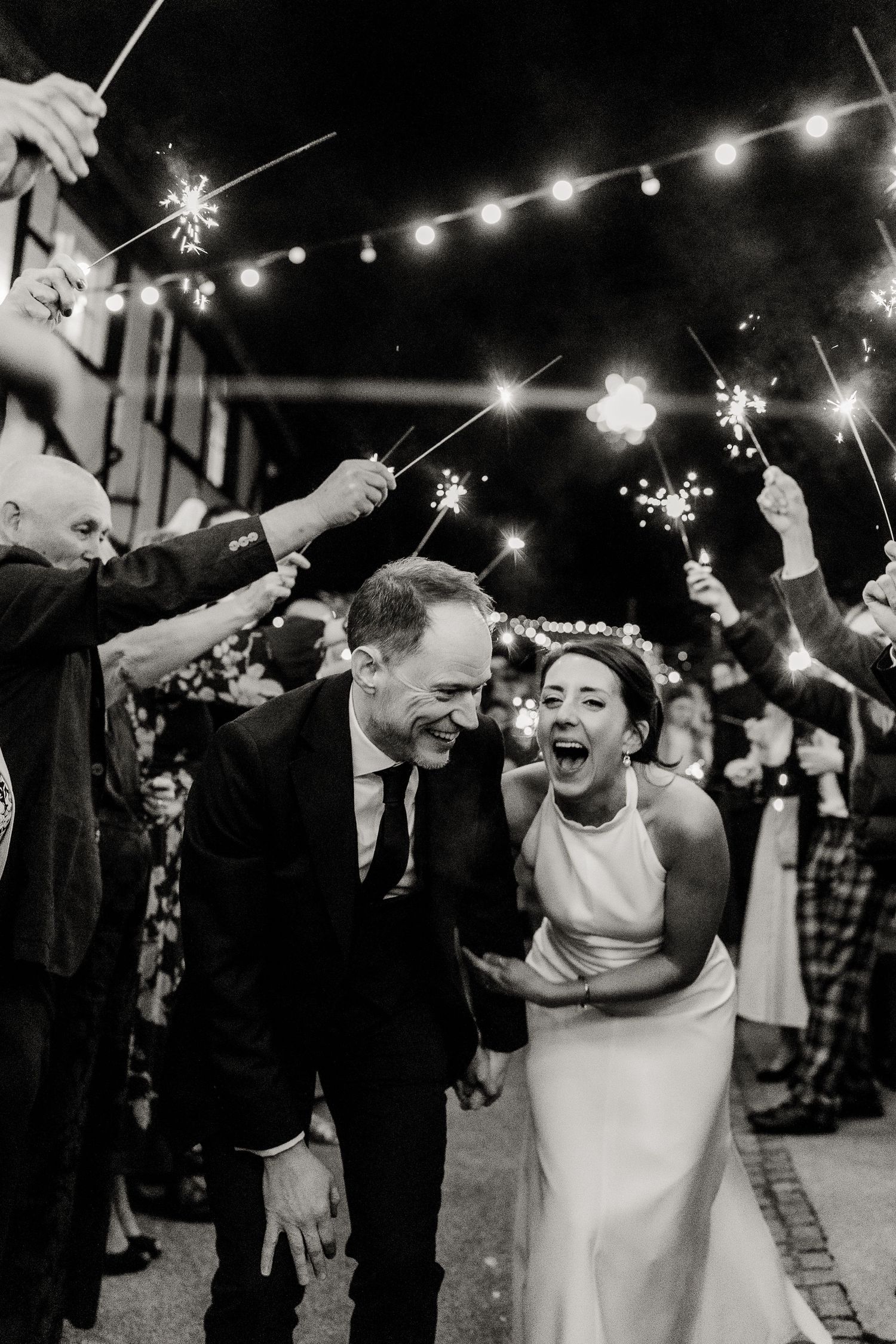 Black and white photo of joyful bride and groom walking through sparkler send-off at night wedding celebration.
