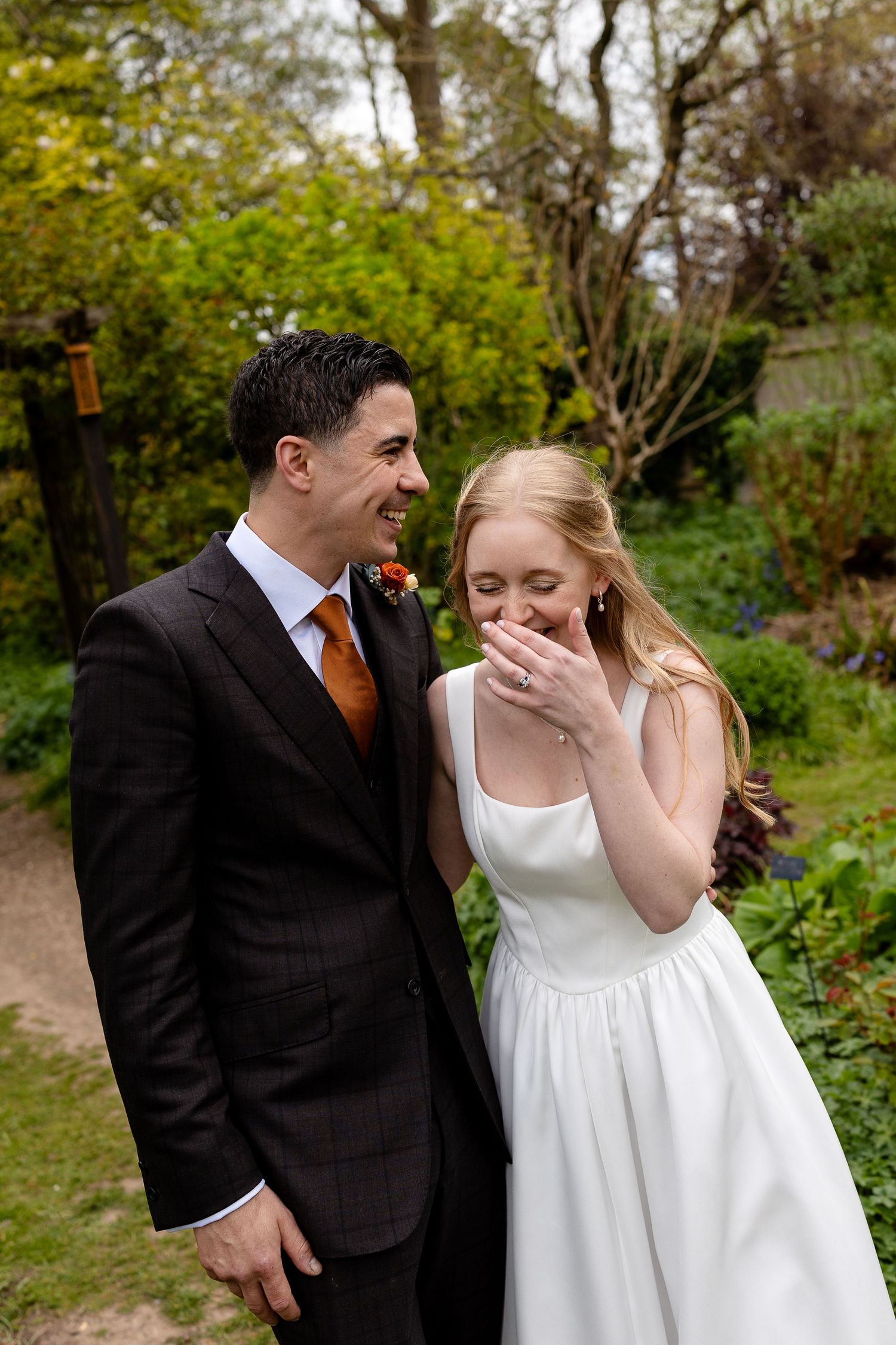 A smiling bride in a white dress and groom in a dark suit with orange tie share a joyful moment in a lush garden.