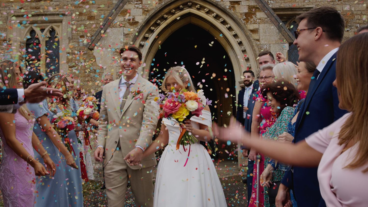Wedding guests shower newlyweds with colorful flower petals as they exit a stone church archway.