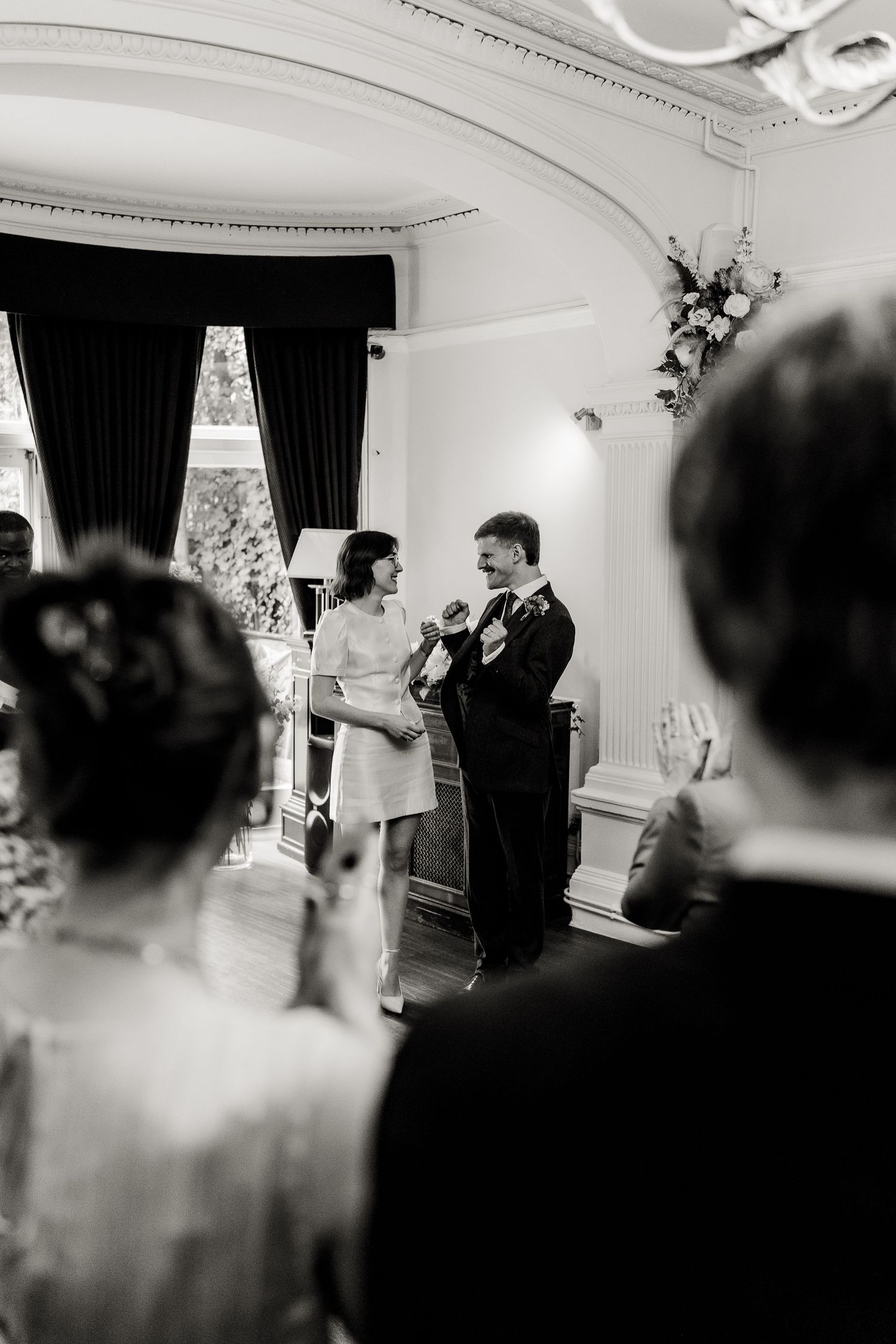 Black and white photo of a bride and groom exchanging vows at an elegant indoor wedding ceremony.