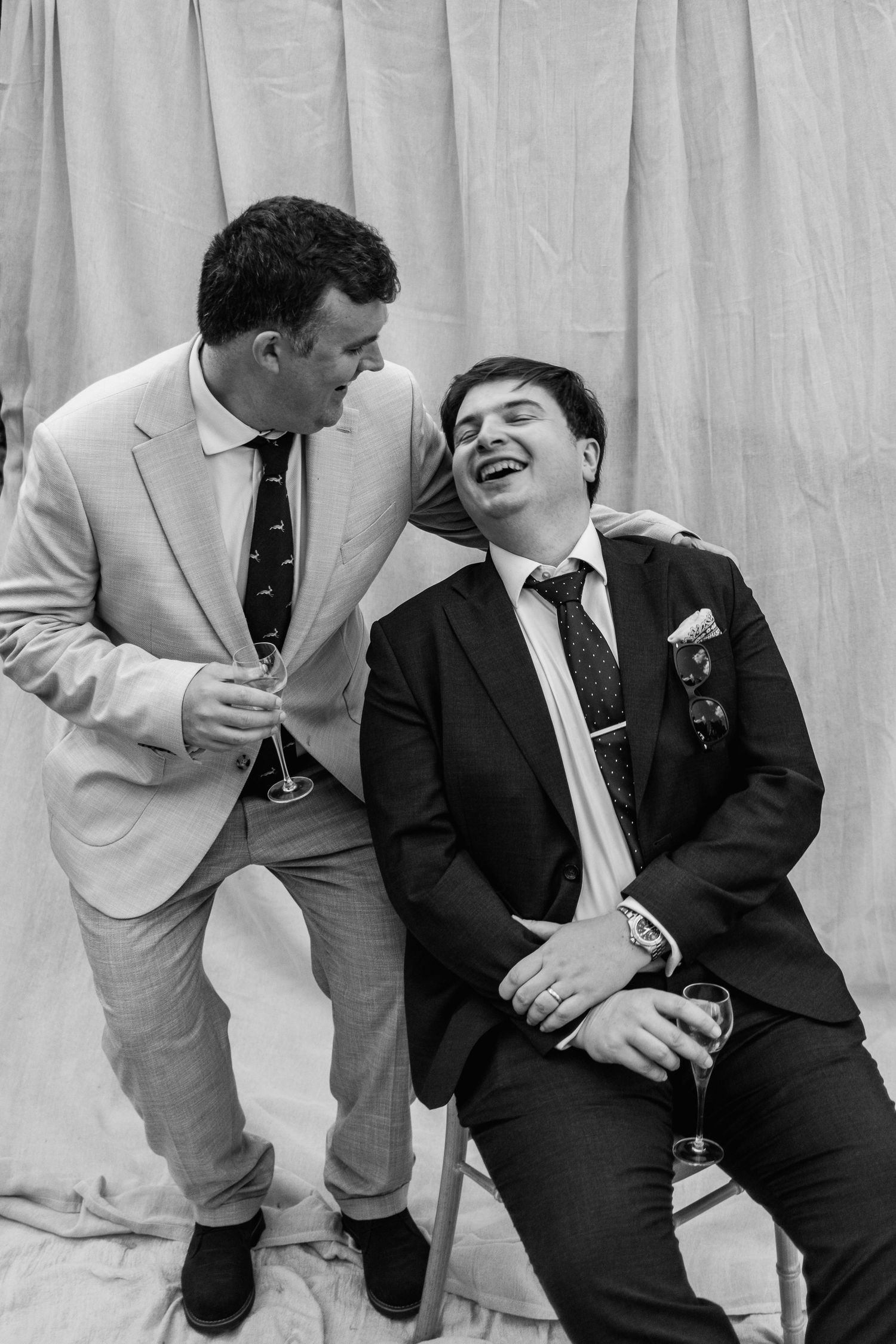 Two men in suits laugh joyfully while holding drinks at a wedding celebration, one seated and one leaning in, in a black and white photo.