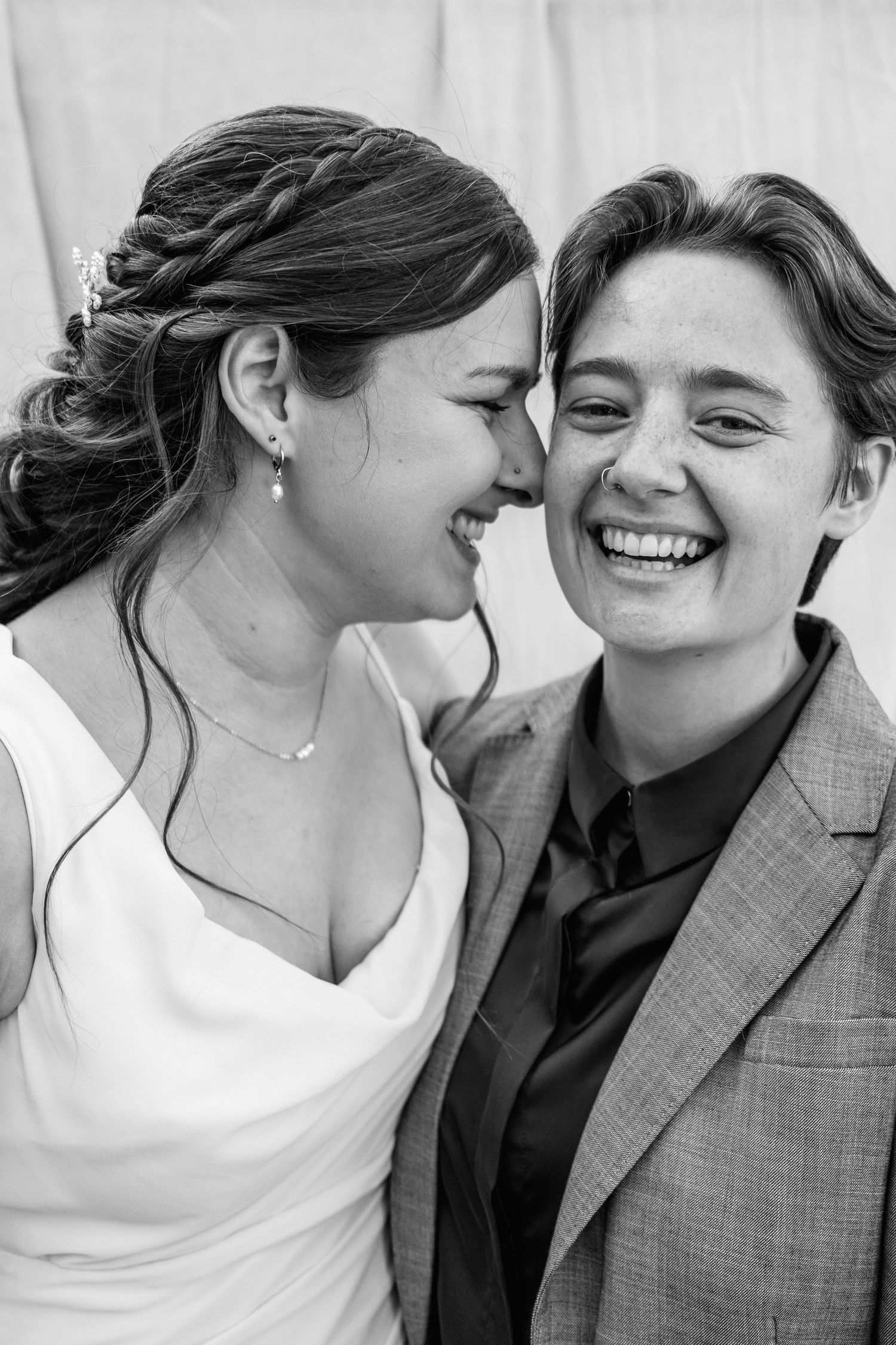 Black and white wedding portrait of two smiling brides laughing together, one in a white gown with a braided updo, the other in a blazer.