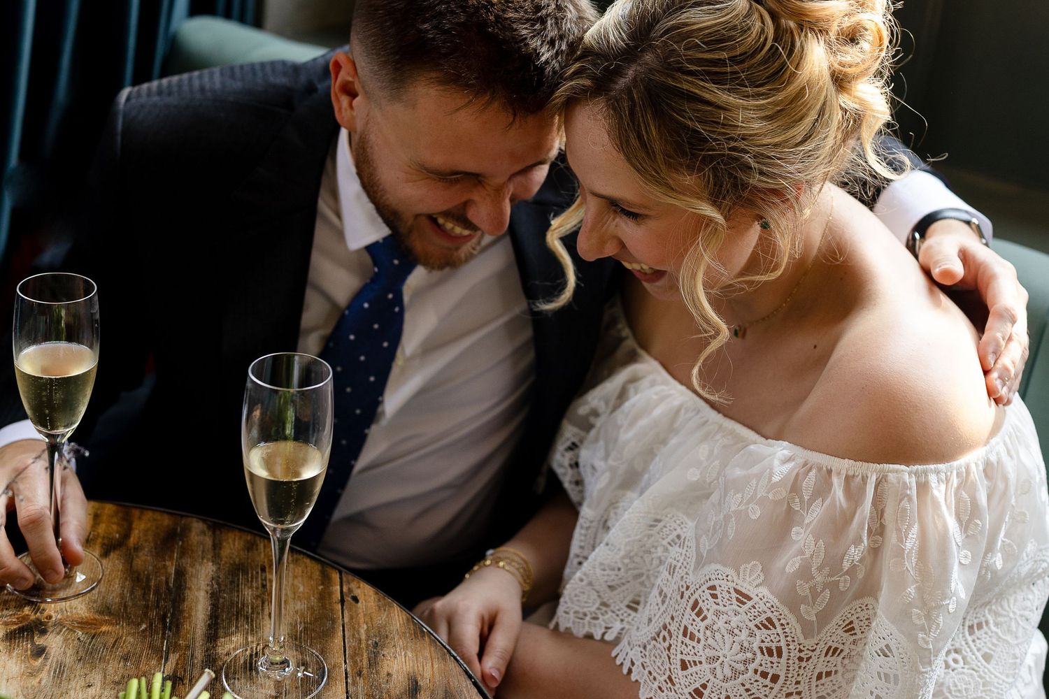 Couple shares intimate moment with champagne glasses at rustic wooden table during wedding celebration.