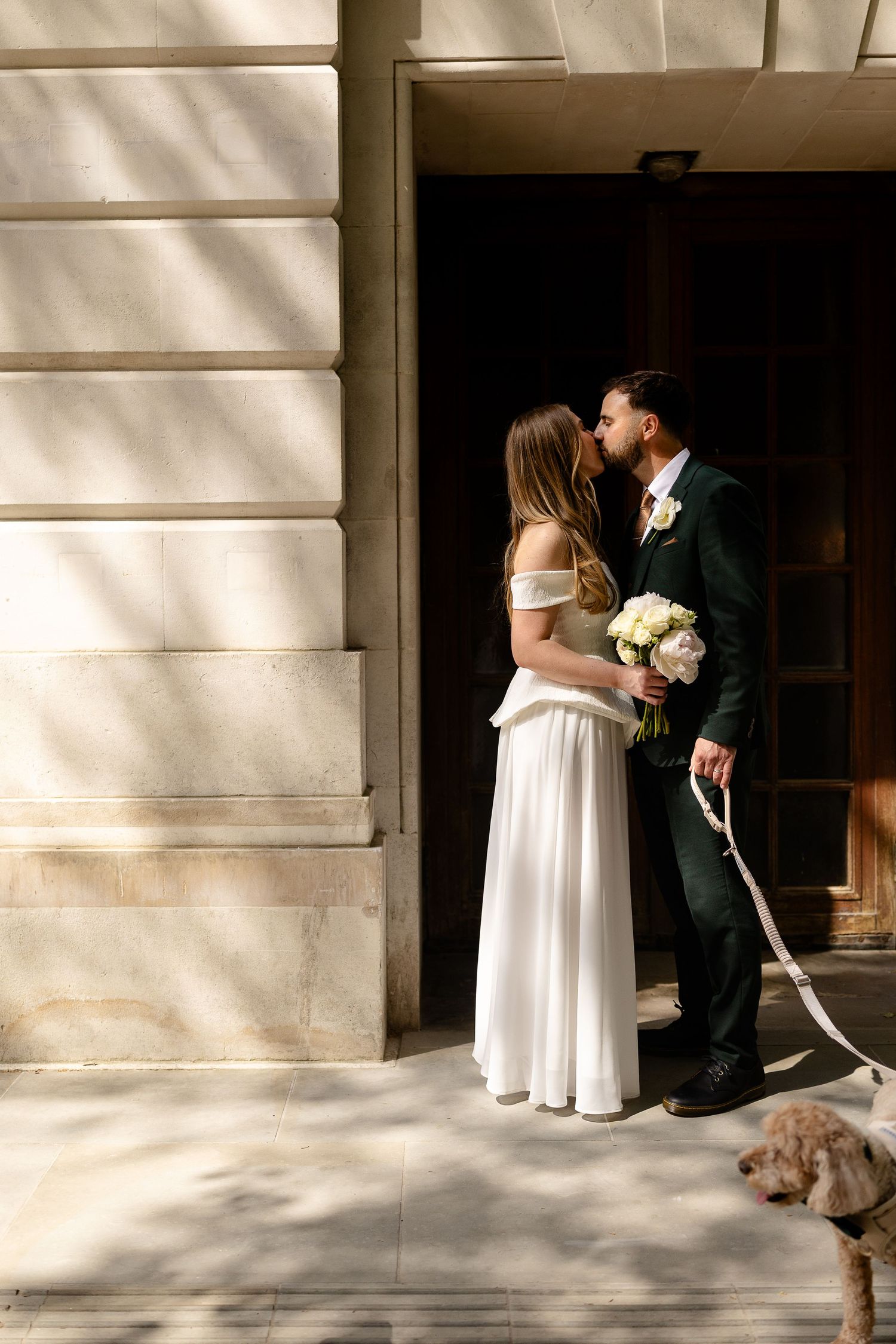 Bride and groom kissing in a sunlit doorway, bride holding white bouquet in elegant off-shoulder gown, groom in dark suit.