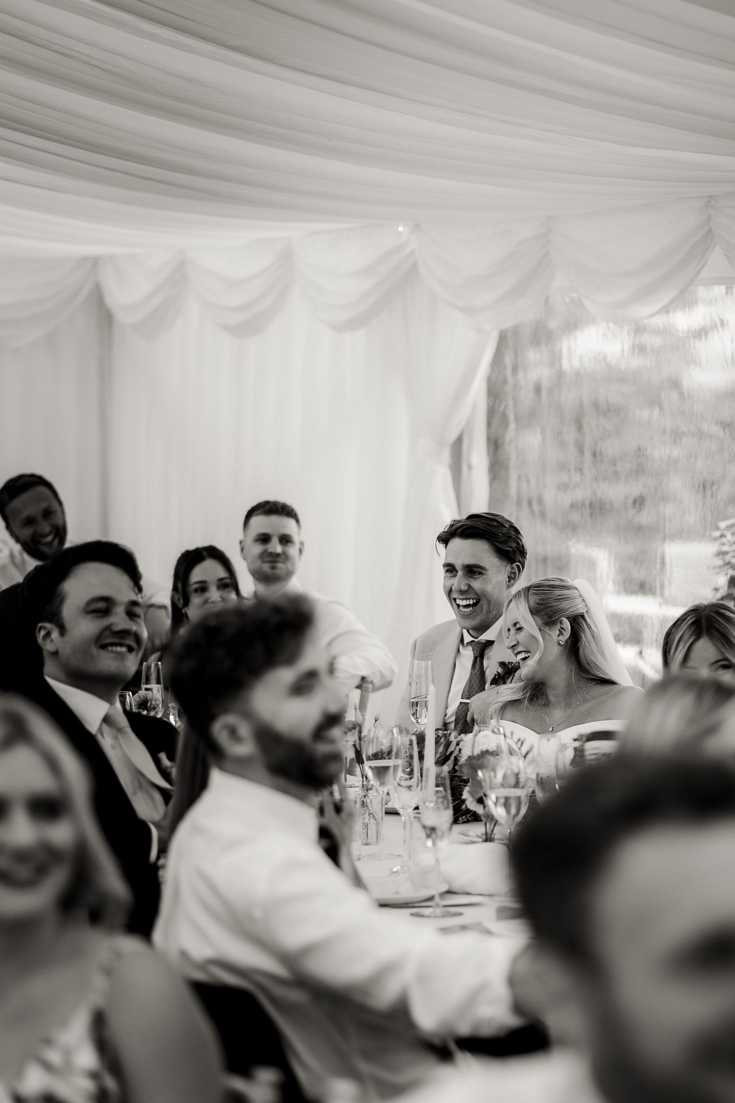 Wedding guests share joyful laughter during reception speeches inside an elegant white tent venue.