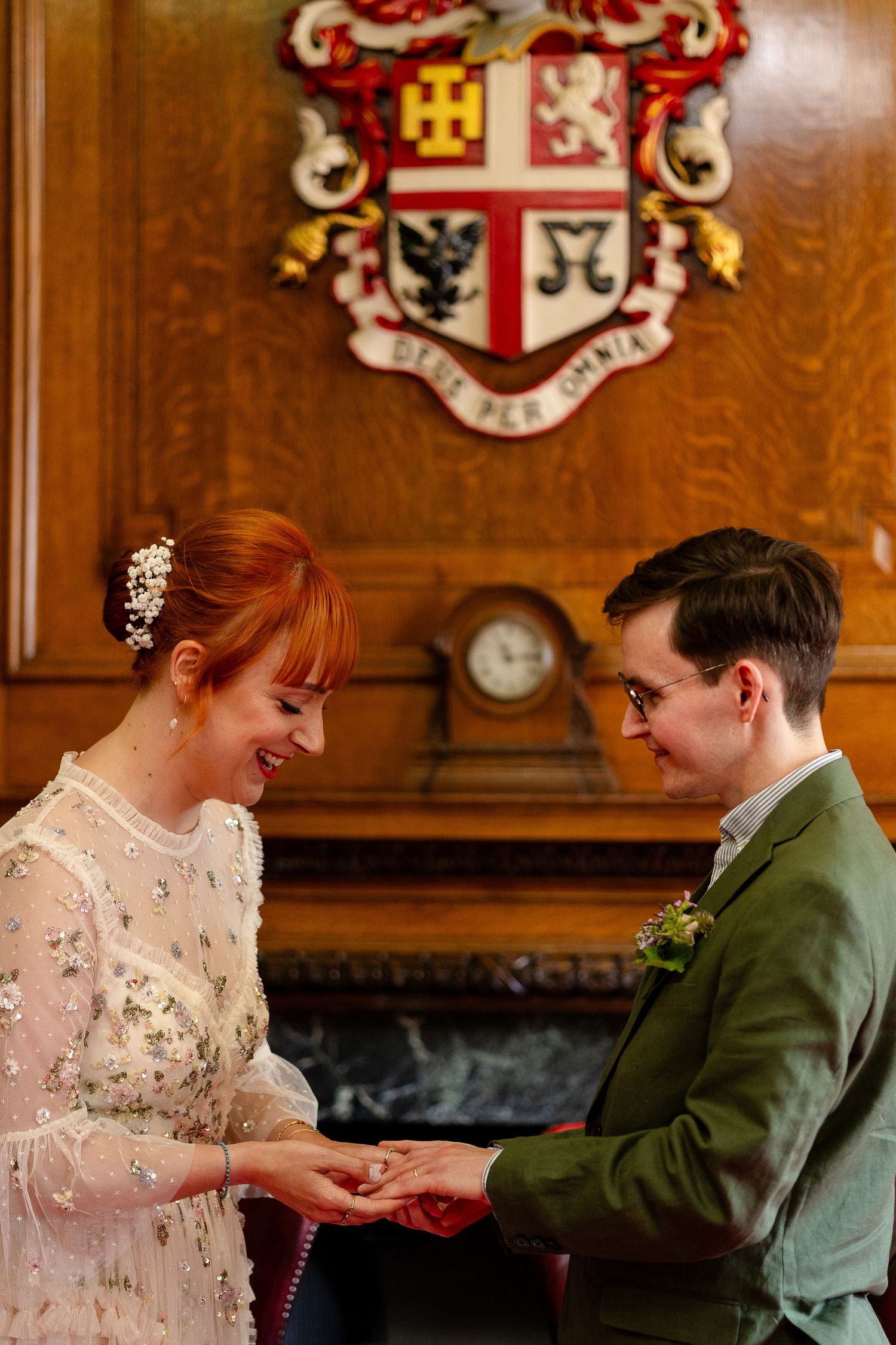 Bride and groom exchange rings at a civil ceremony beneath a wooden coat of arms in an elegant registry office.
