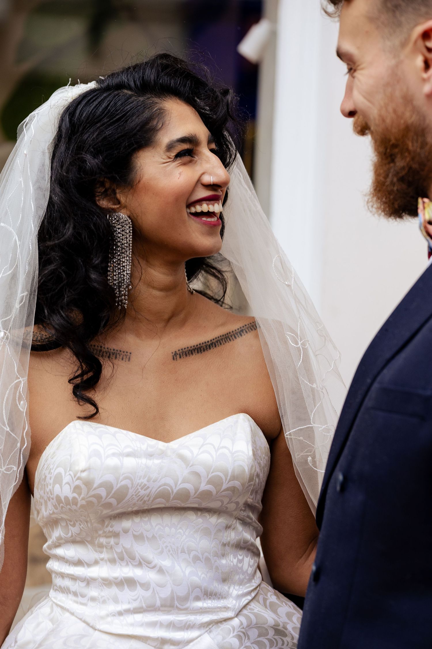Bride in white strapless gown and veil laughs joyfully at bearded groom in navy suit during wedding ceremony.