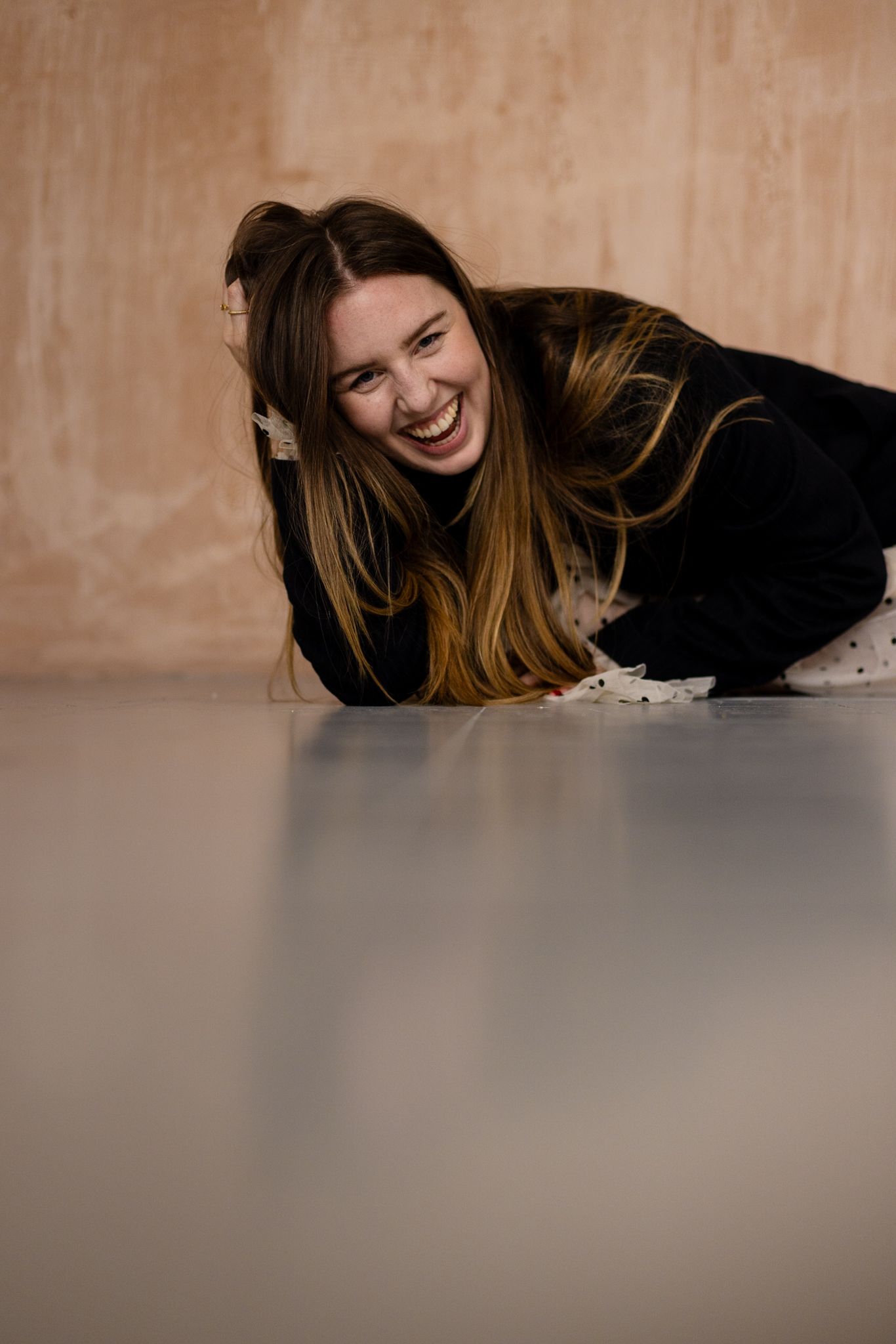 A joyful person with long blonde hair wearing a black sweater lies on a smooth floor, laughing heartily against a beige wall.